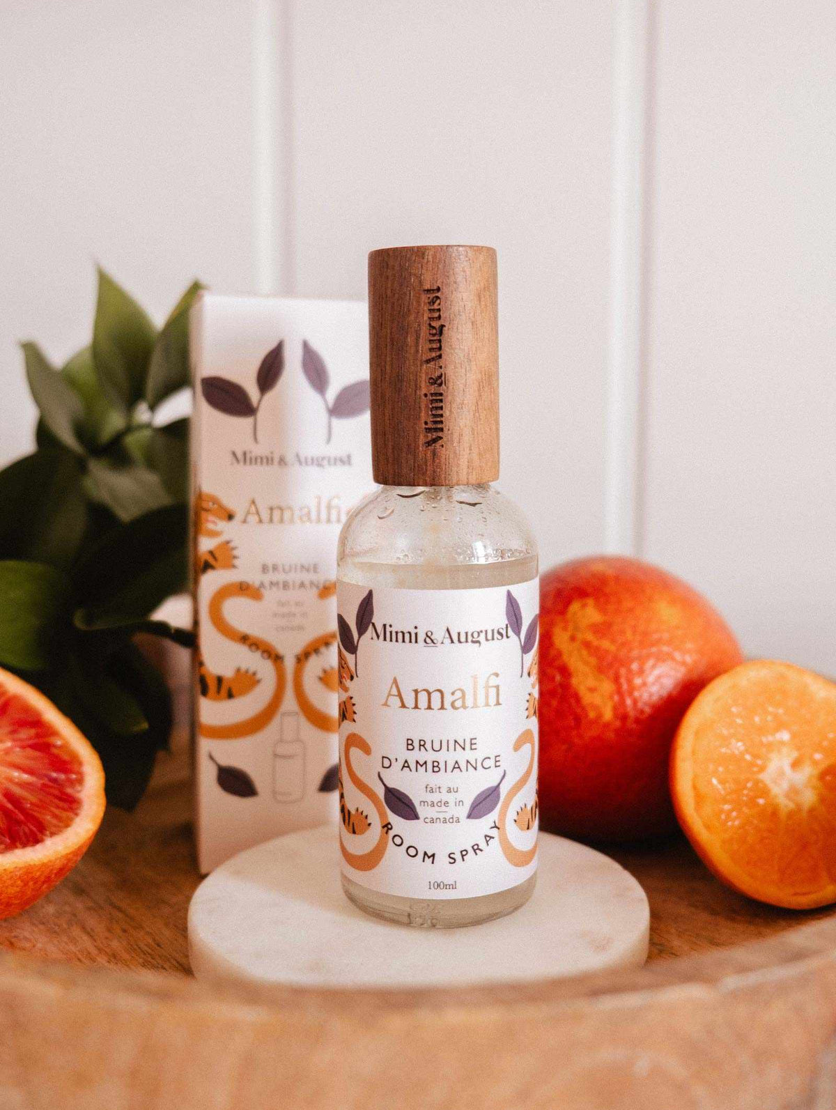 The Mimi & August Room spray - Amalfi sits on a wooden tray with its packaging, accented by orange halves and green leaves, evoking a Mediterranean vibe with its refreshing citrus aroma.