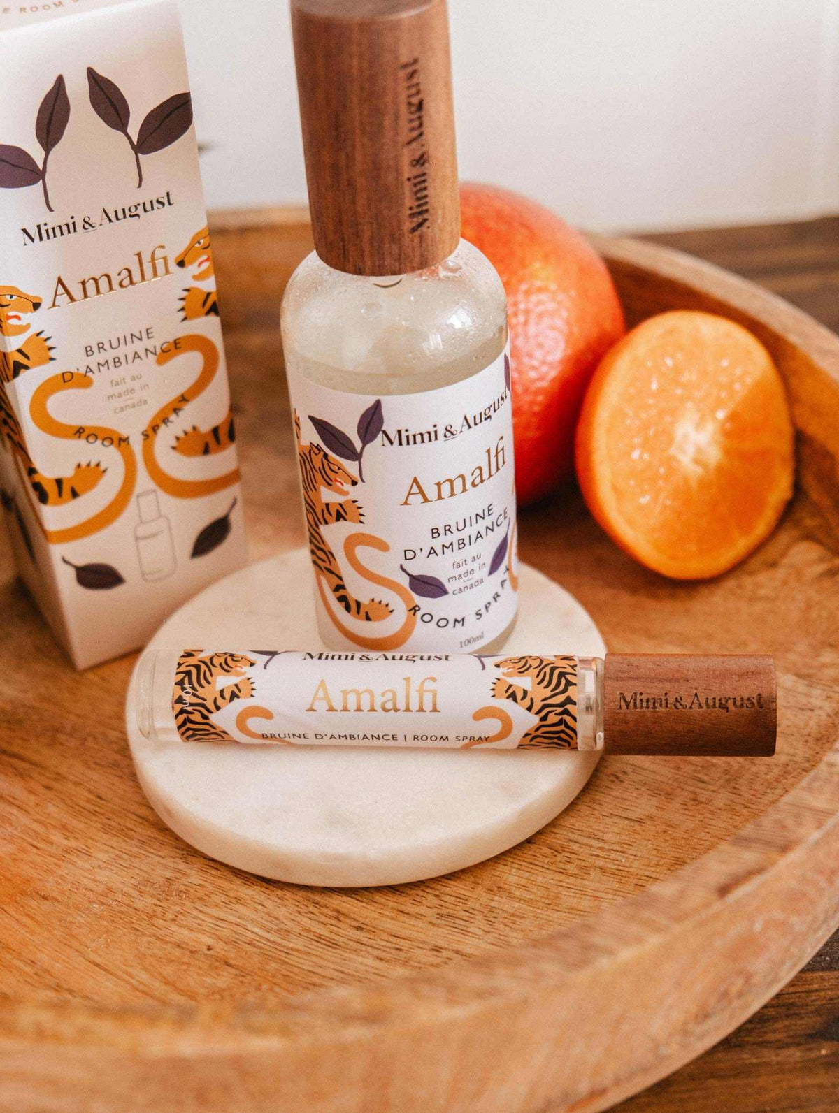 A bottle and spray of Mimi & August Room Spray - Amalfi with packaging rest on a wooden tray next to a whole orange and an orange half, evoking the fresh citrus aroma of the Mediterranean.