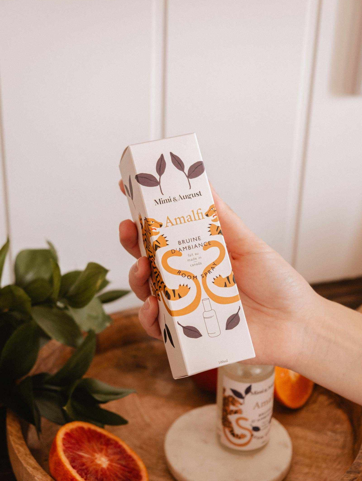 A hand holds a boxed Room Spray - Amalfi by Mimi & August with a tiger design. Nearby, on a wooden tray with leaves and an orange, the same spray evokes a Mediterranean escape and citrusy freshness.