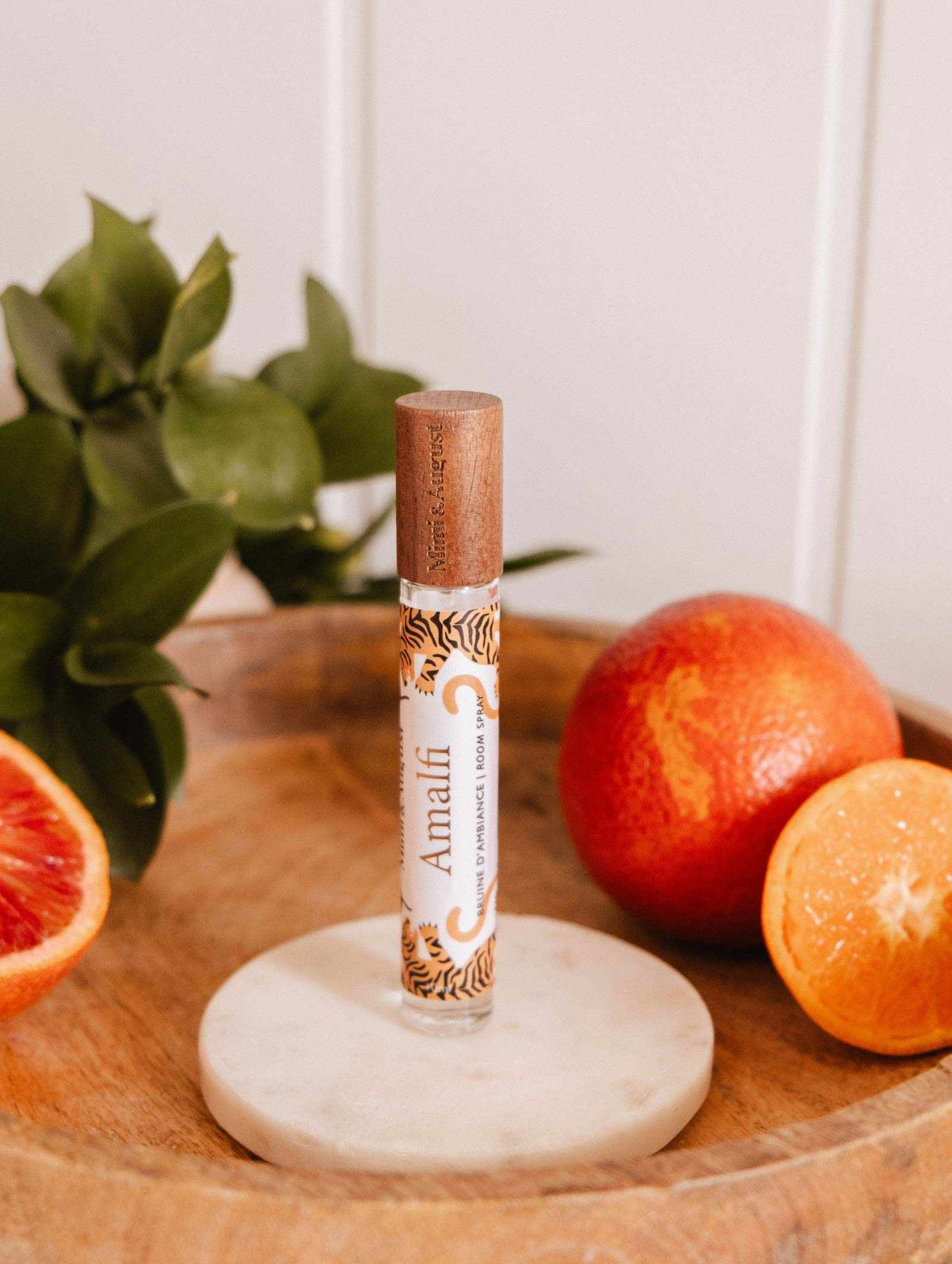 A bottle of Mimi & August Room Spray - Amalfi stands upright on a round marble coaster with blood oranges and green leaves, evoking the Mediterranean fragrance and citrus sparkle of this Amalfi-inspired scent.