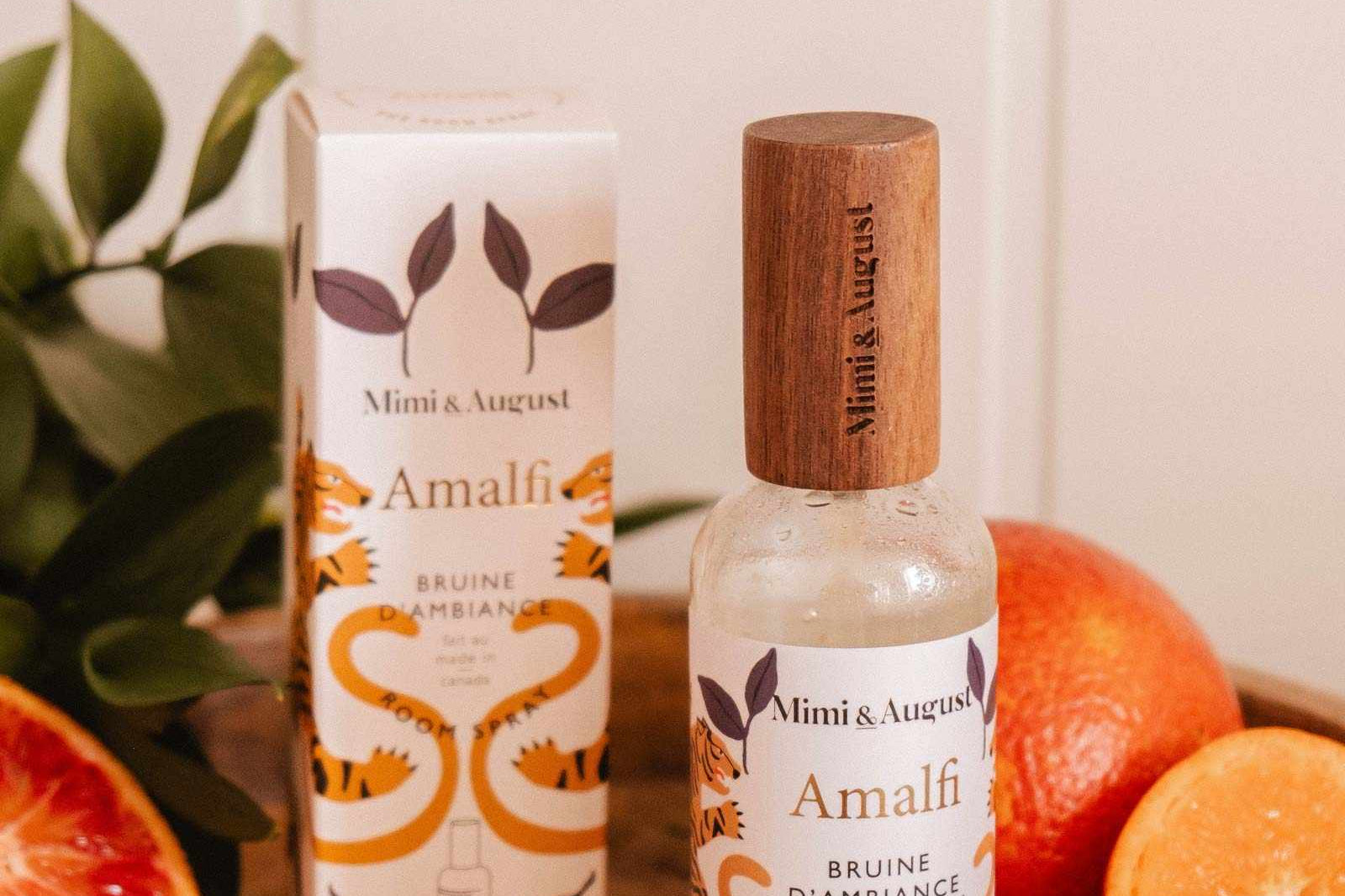 A bottle and box of Mimi & August Room spray - Amalfi are shown on a wooden surface with cut oranges and green leaves, evoking a refreshing citrus scent for a Mediterranean-inspired space.