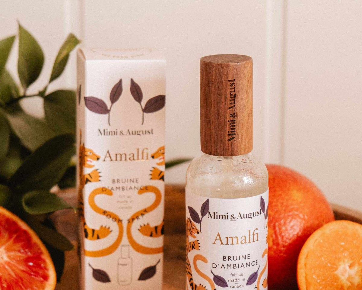 A bottle and box of Mimi & August Room spray - Amalfi are shown on a wooden surface with cut oranges and green leaves, evoking a refreshing citrus scent for a Mediterranean-inspired space.