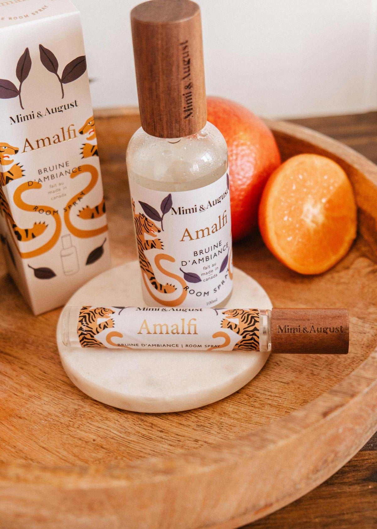 Une bouteille et un vaporisateur de bruine d'ambiance Amalfi de Mimi & August avec son emballage reposent sur un plateau en bois à côté d'une orange entière et d'une moitié d'orange, évoquant l'arôme frais d'agrumes de la Méditerranée.