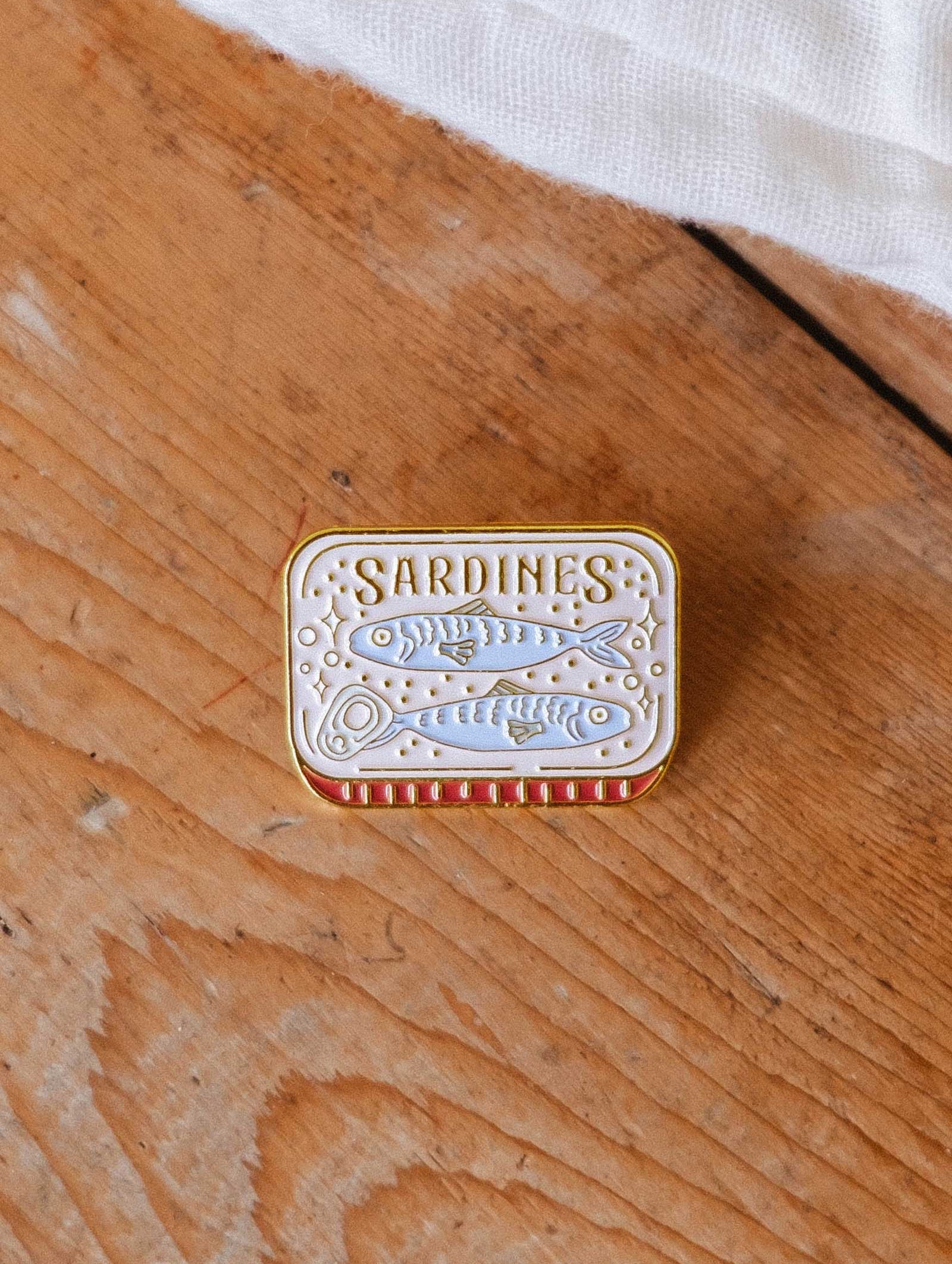 A small, rectangular Cozy Sardines Enamel Pin by Mimi & August sits on a wooden surface. Designed in Montreal, it features a sardine illustration and a red and white striped border.