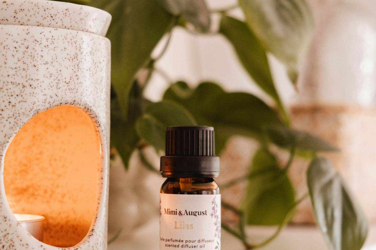 A small bottle labeled "Mimi & August Scented Oil - Lilac" is placed on a shelf beside a white ceramic oil diffuser with a lit candle inside, releasing its calming and soothing lilac scent. In the background, green leaves and neutral decor are visible.