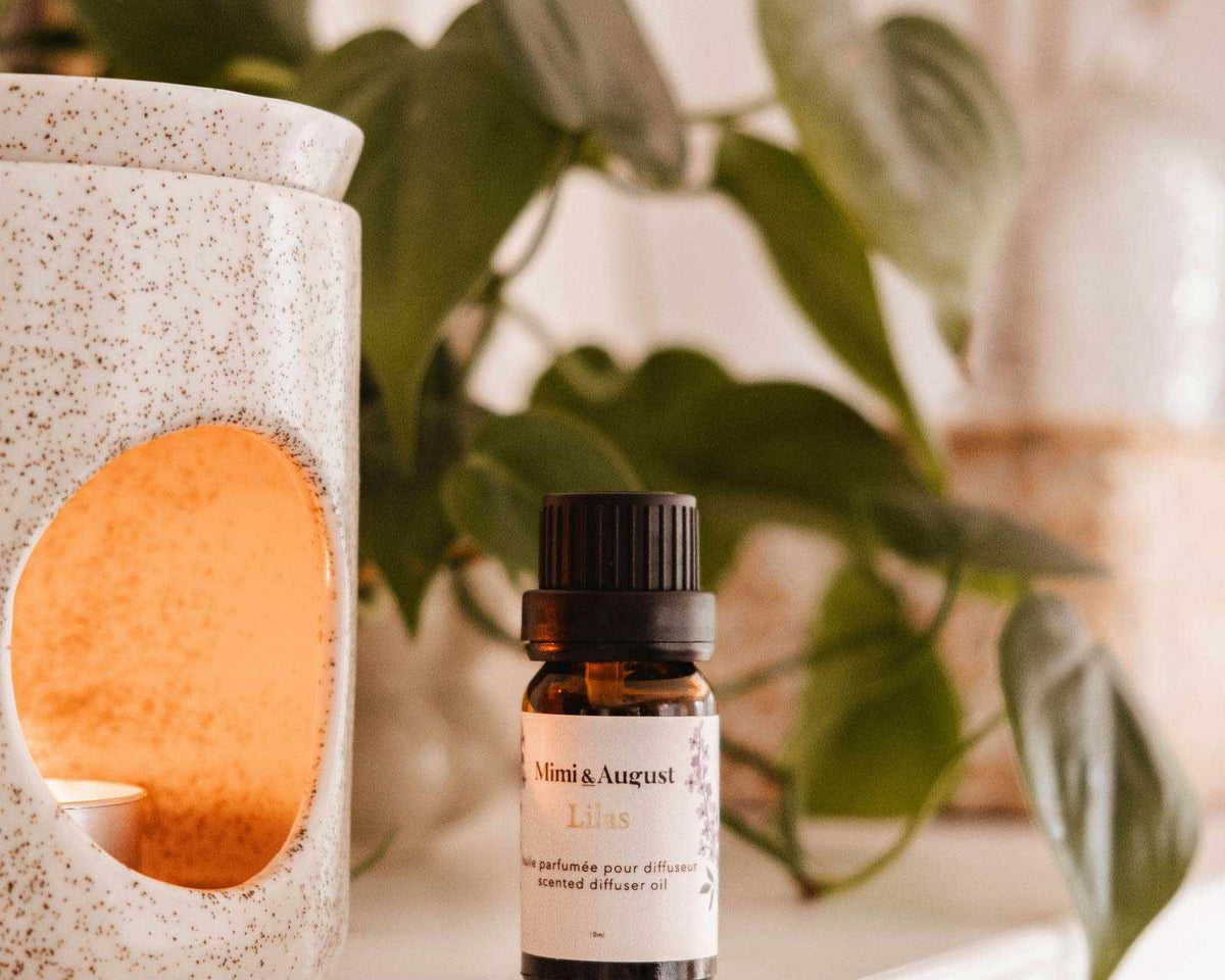 A small bottle labeled "Mimi & August Scented Oil - Lilac" is placed on a shelf beside a white ceramic oil diffuser with a lit candle inside, releasing its calming and soothing lilac scent. In the background, green leaves and neutral decor are visible.