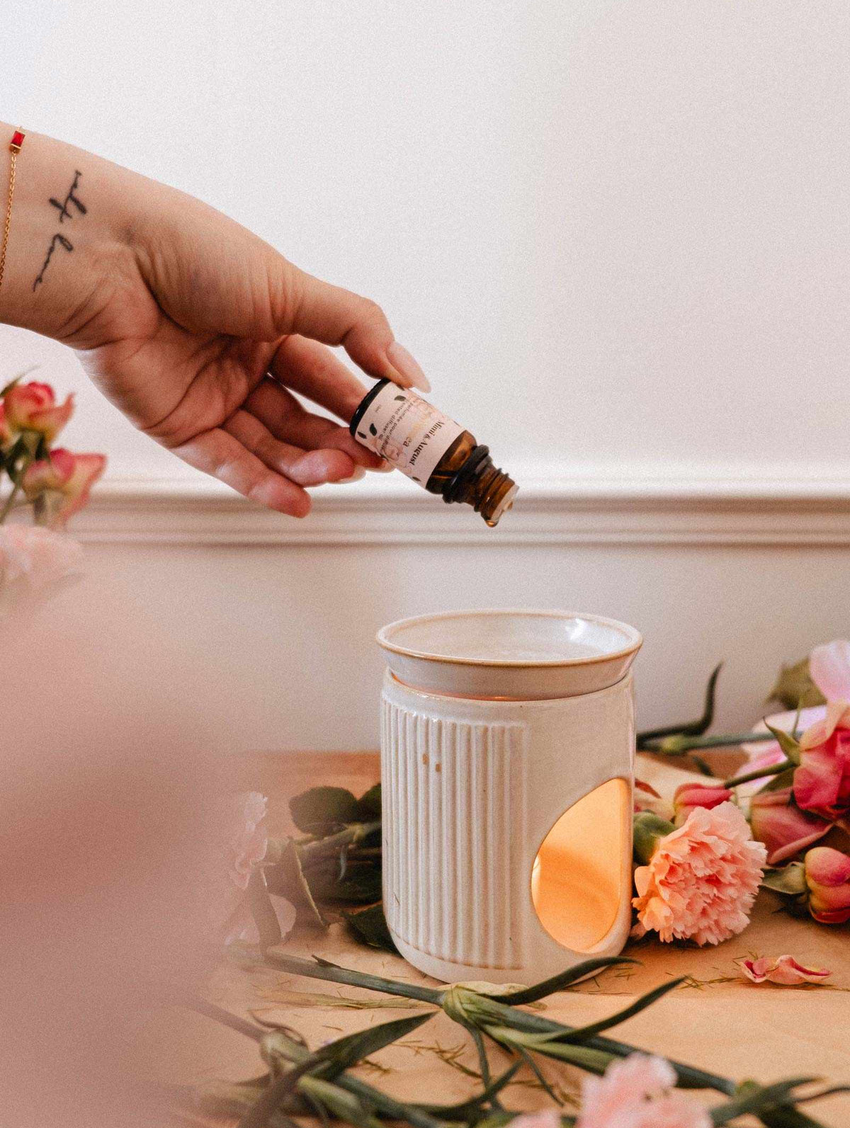 Une main verse l'huile parfumée Musea de Mimi & August, au parfum d'orange sanguine, dans un brûleur en céramique blanc entouré de fleurs roses — une charmante alternative au diffuseur ultrasonique.