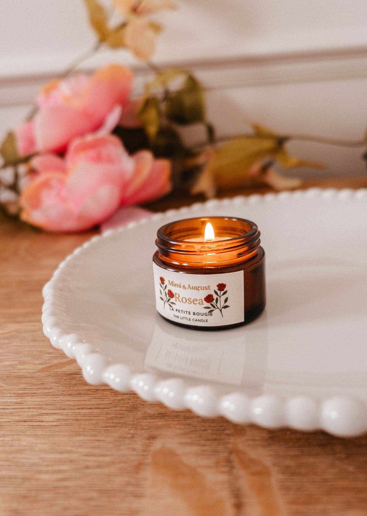 La bougie de soya Rosea 2 oz de Mimi & August, dans un pot en verre brun, est allumée et posée sur une assiette blanche festonnée avec des fleurs artificielles roses en arrière-plan sur une surface en bois.