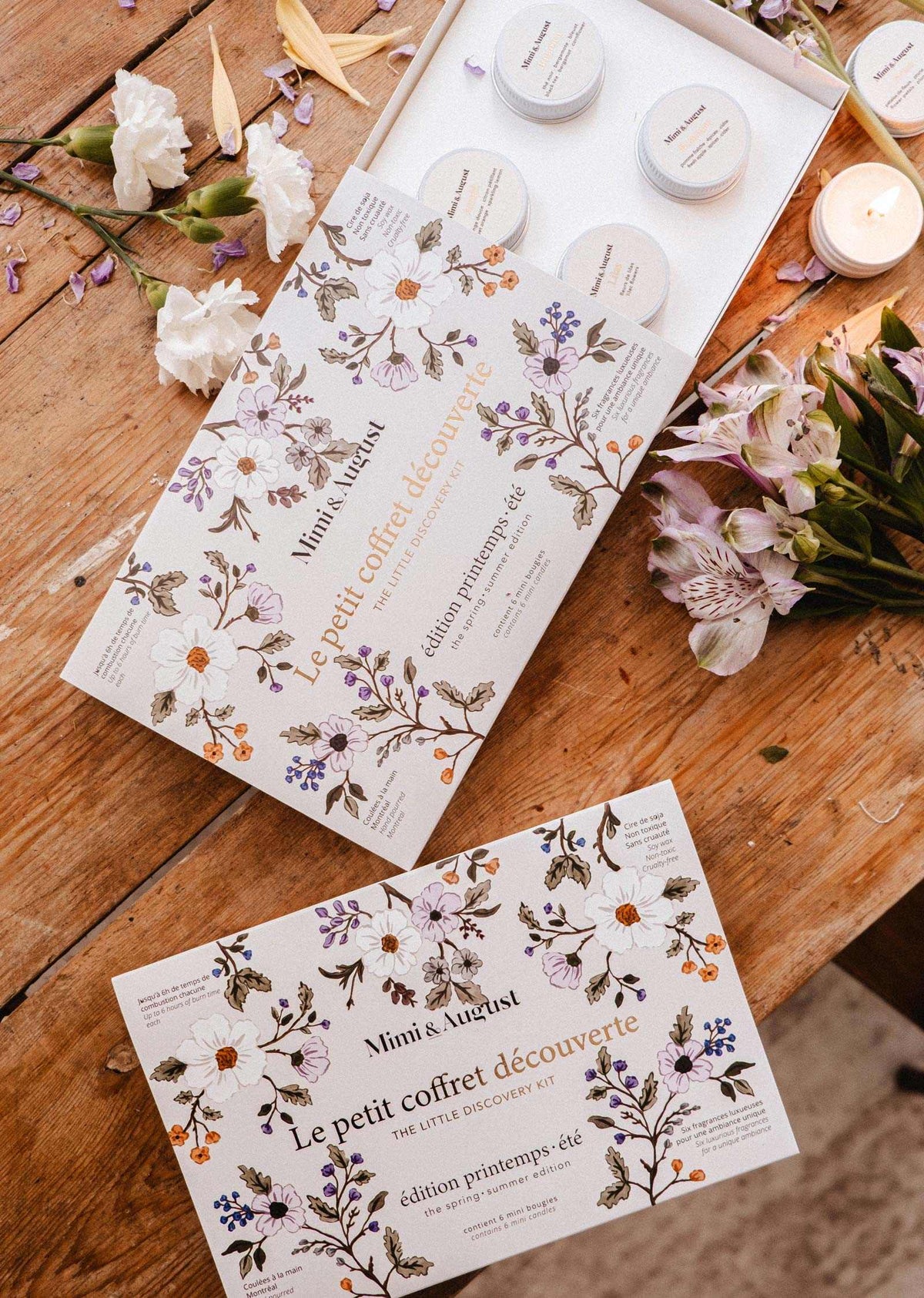 Le Kit Découverte Printemps/Été de Mimi & August et un mini lampion en cire de soya sont présentés sur une table en bois avec des fleurs blanches et violettes éparpillées.