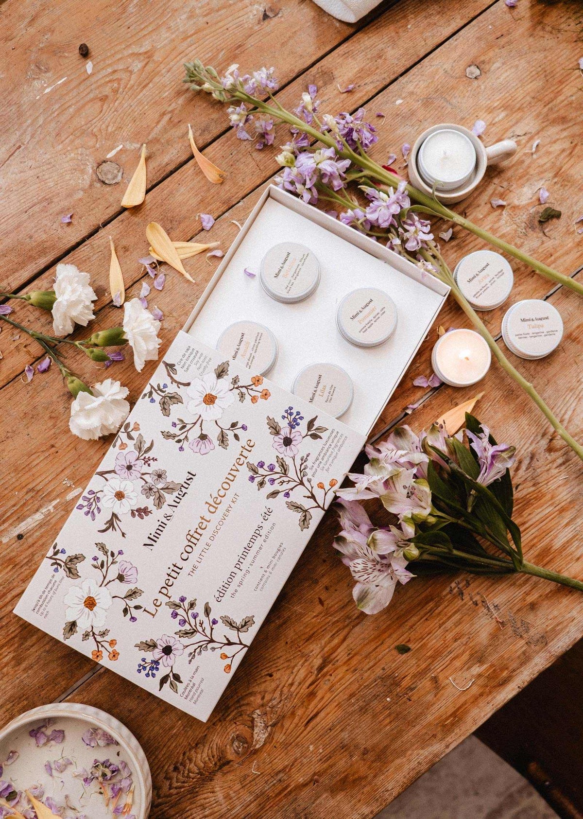 Le Kit Découverte Printemps/Été de Mimi & August : un coffret cadeau ouvert au thème floral avec de petits pots, un mini lampion en cire de soya et un coffret de lampions, joliment disposés parmi des fleurs et des bougies sur une table en bois rustique.