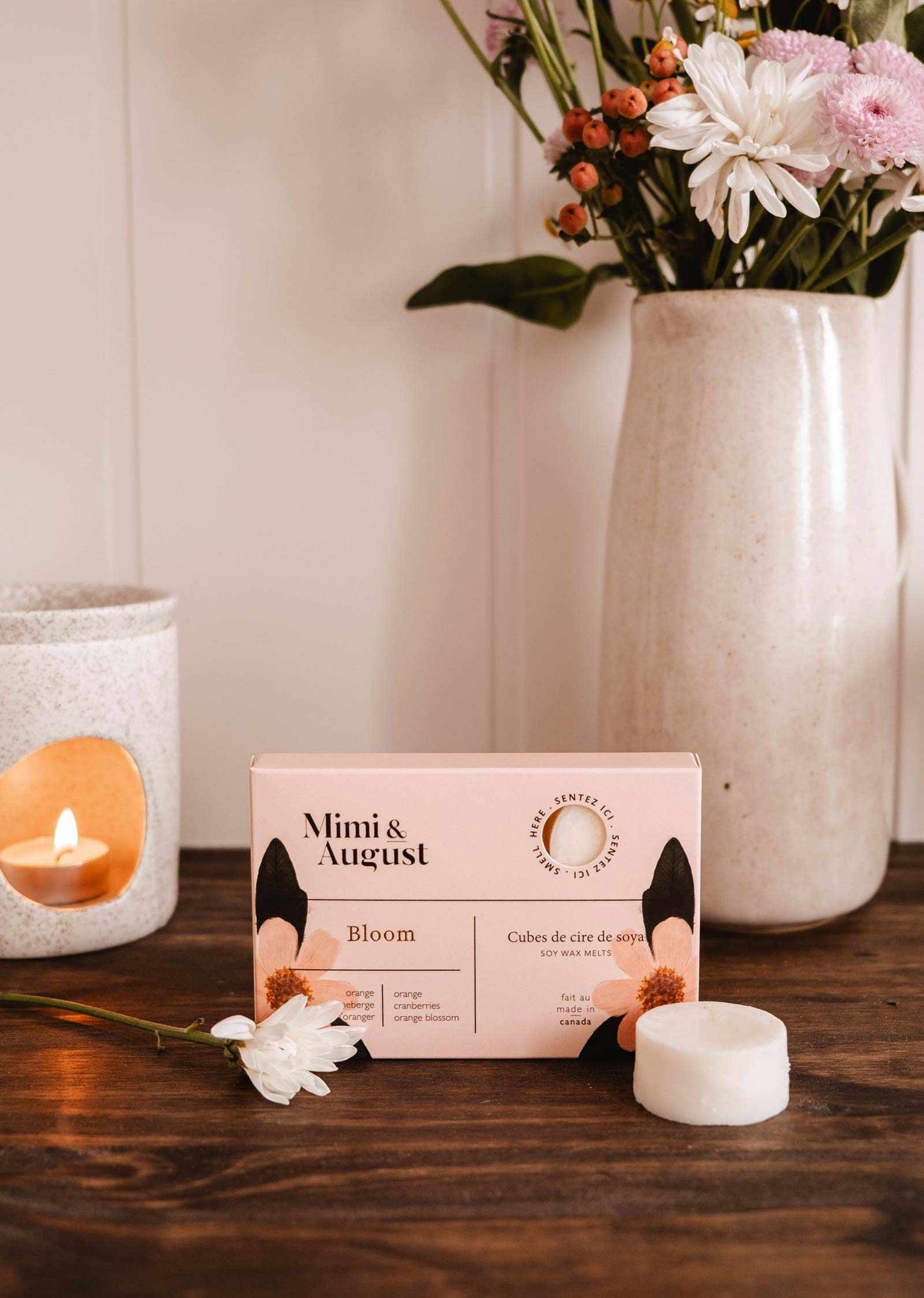 Sur une table en bois, entourée de fleurs, d'une petite bougie blanche et d'un vase de fleurs variées, repose une boîte de fondants de cire de soya Bloom de Mimi & August. Ces cubes écoresponsables offrent un parfum longue durée pour toute pièce.