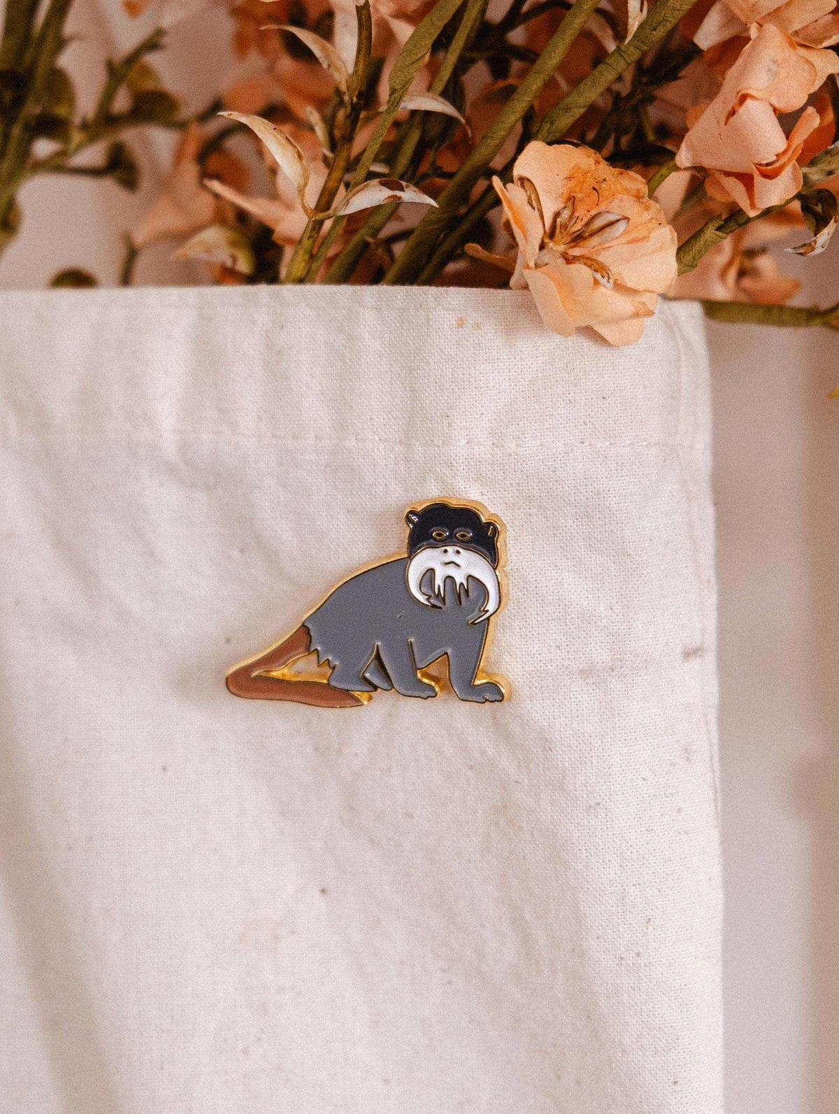 Épinglette Tamarin de Mimi & August est un accessoire plein de fantaisie pour les amoureux des animaux. Elle met en vedette un petit singe au joli moustache blanc, posé sur un fond de tissu blanc, accompagné de fleurs séchées