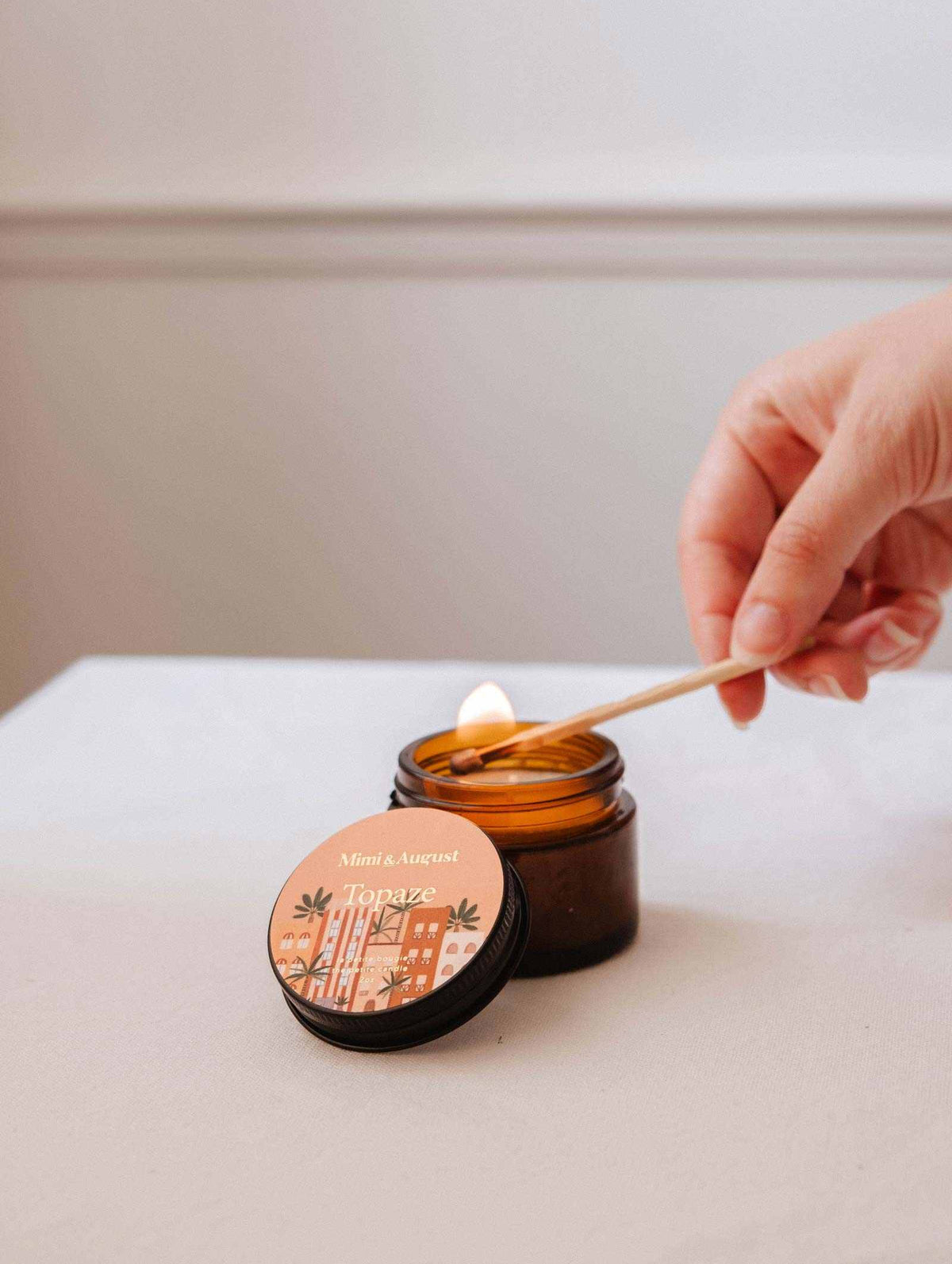 Une main allume la bougie de soya Topaze de 2 oz de Mimi & August dans un pot en verre brun avec une allumette; son couvercle repose à proximité — une bougie réutilisable idéale pour ajouter un parfum chaleureux de bois de santal et de vanille à votre maison.