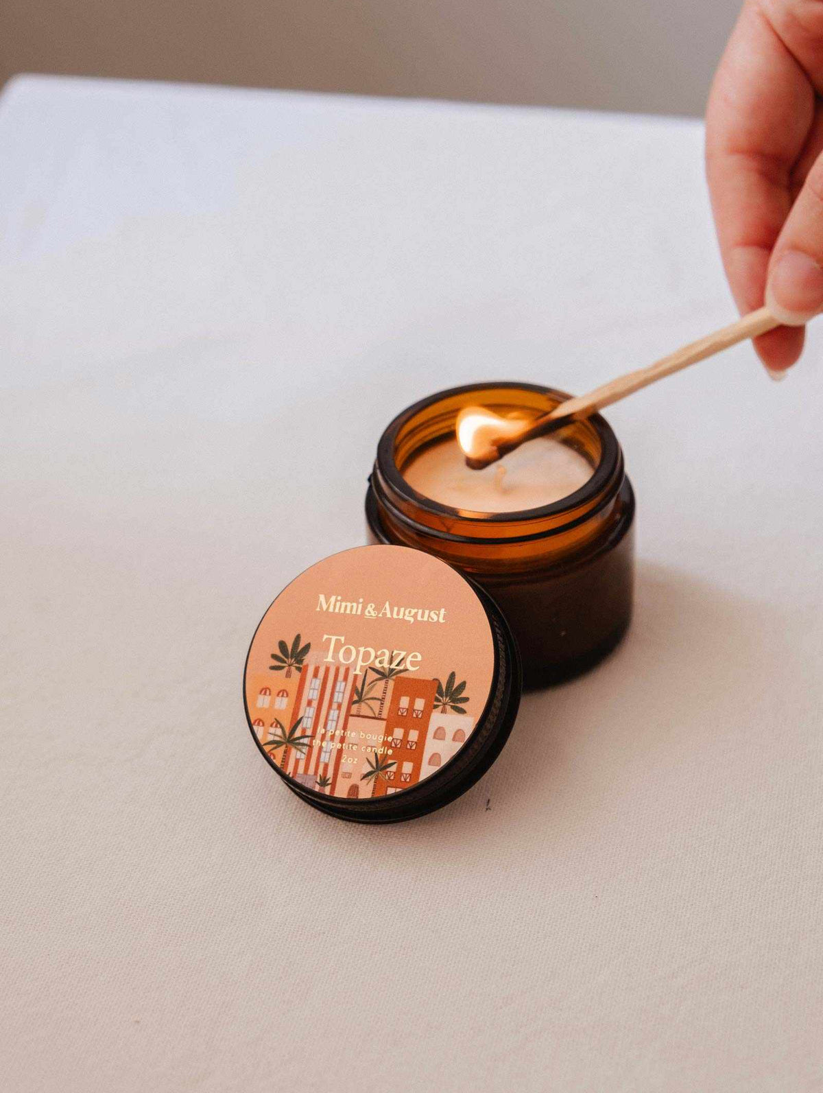 Une main allume la bougie de soya Topaze de 2 oz de Mimi & August dans un pot en verre brun sur une surface beige, emplissant votre espace d'un parfum de bois de santal et de vanille.