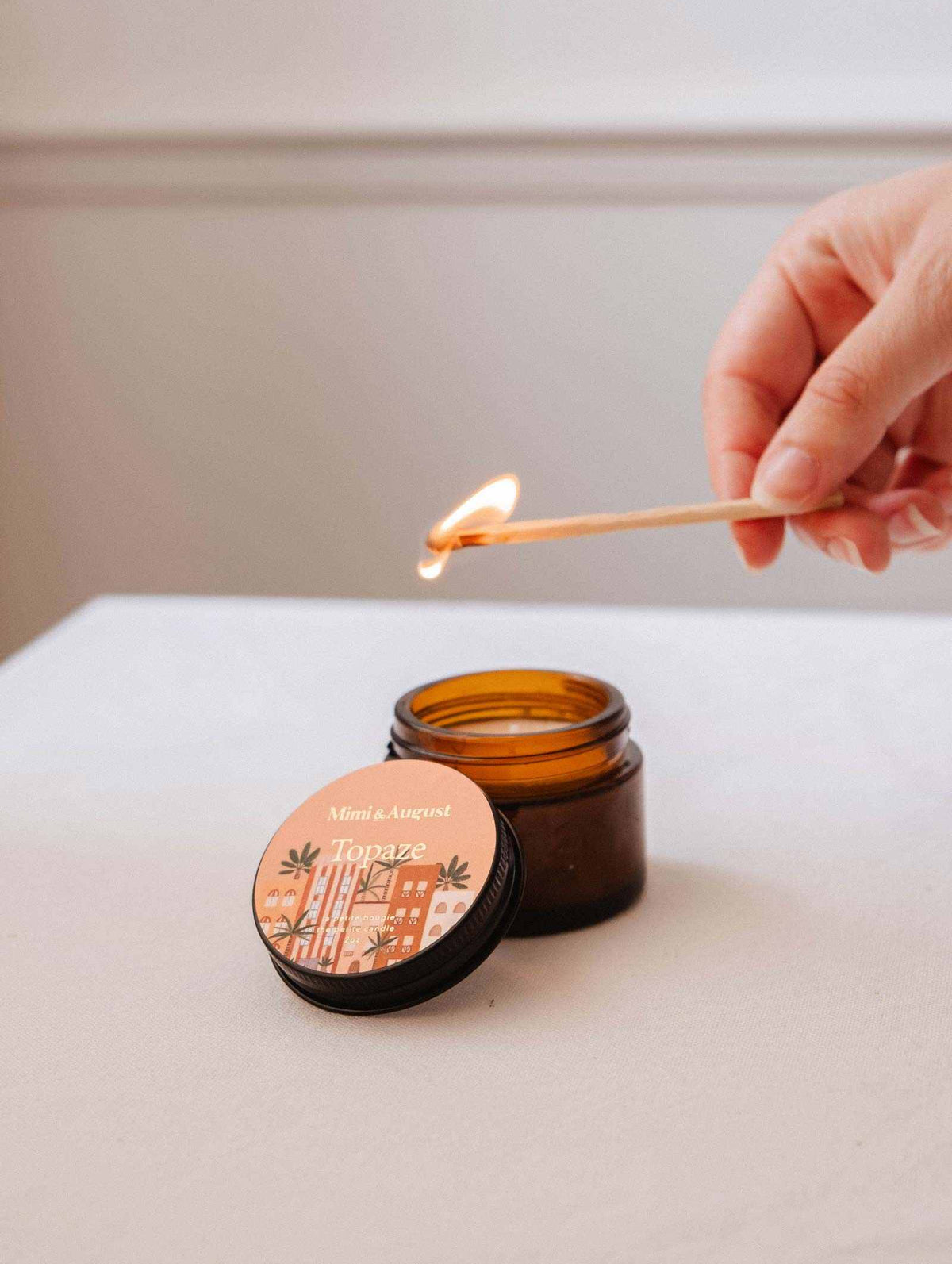 Une main allume une allumette en bois au-dessus d'une bougie de soya Topaze de 2 oz de Mimi & August ouverte sur une surface blanche, libérant un chaleureux parfum d'ambiance de bois de santal et de vanille.