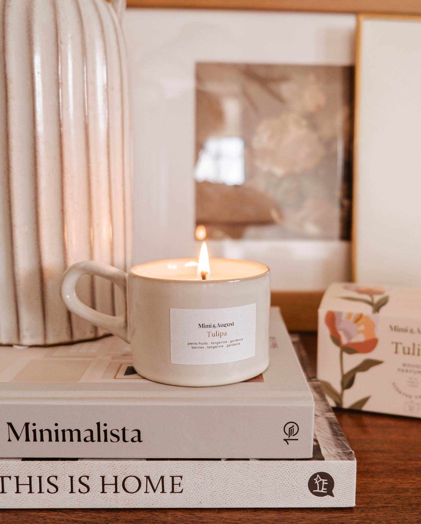 A Mimi & August Tulipa reusable candle on top of a stack of books titled "minimalista" and "this is home," next to a framed photo and a vase.