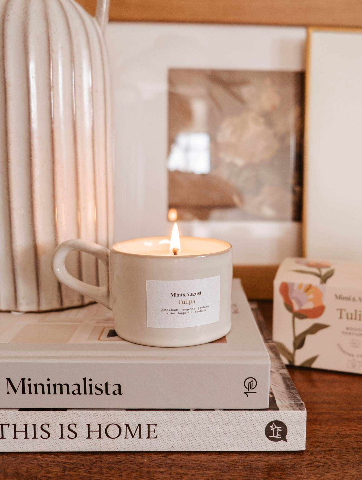 A Mimi & August Tulipa reusable candle on top of a stack of books titled "minimalista" and "this is home," next to a framed photo and a vase.