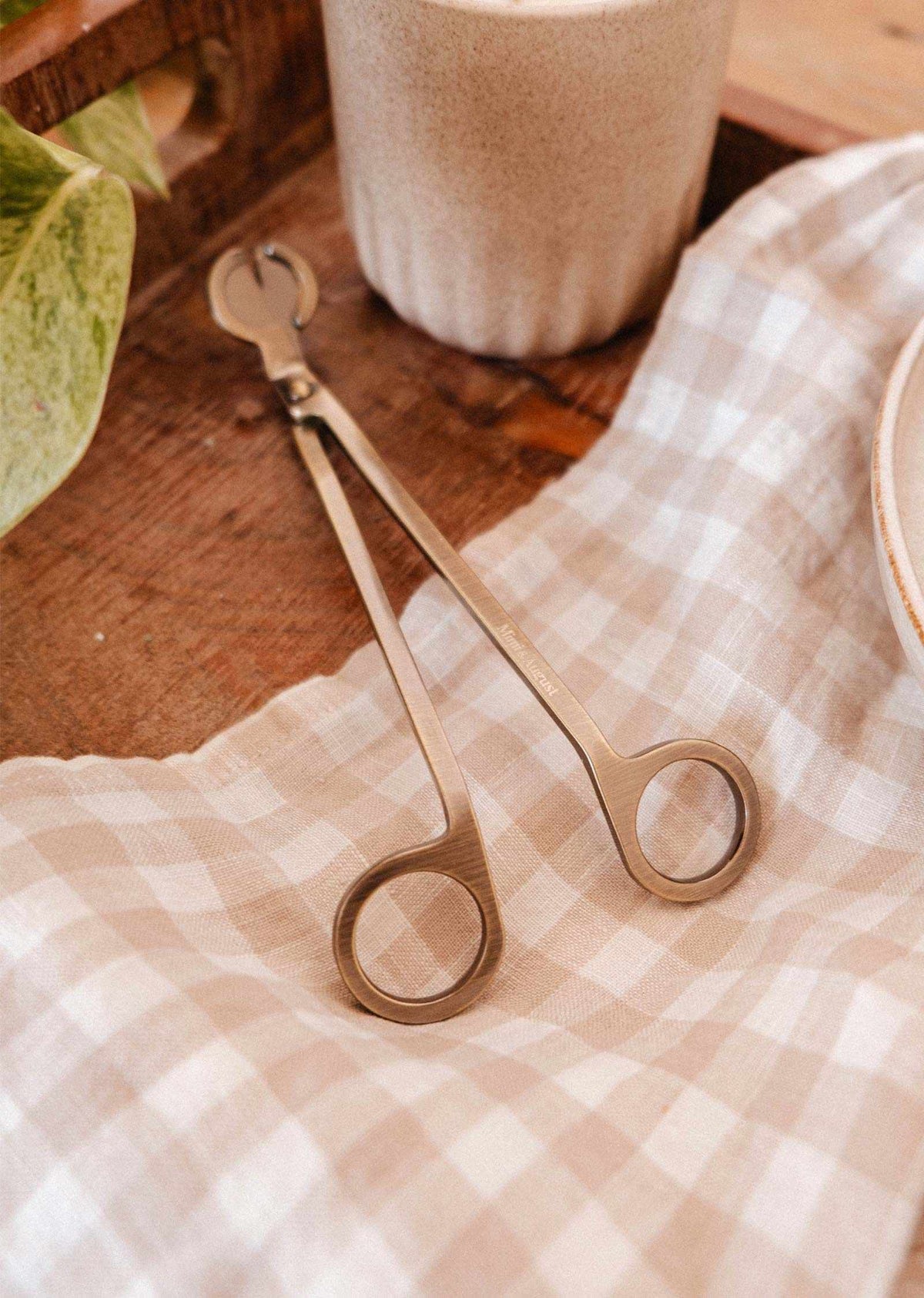 The Mimi & August Wick Trimmer, made of stainless steel, rests on a beige checkered cloth beside a candle and plant, creating an elegant candle accessory for your home.