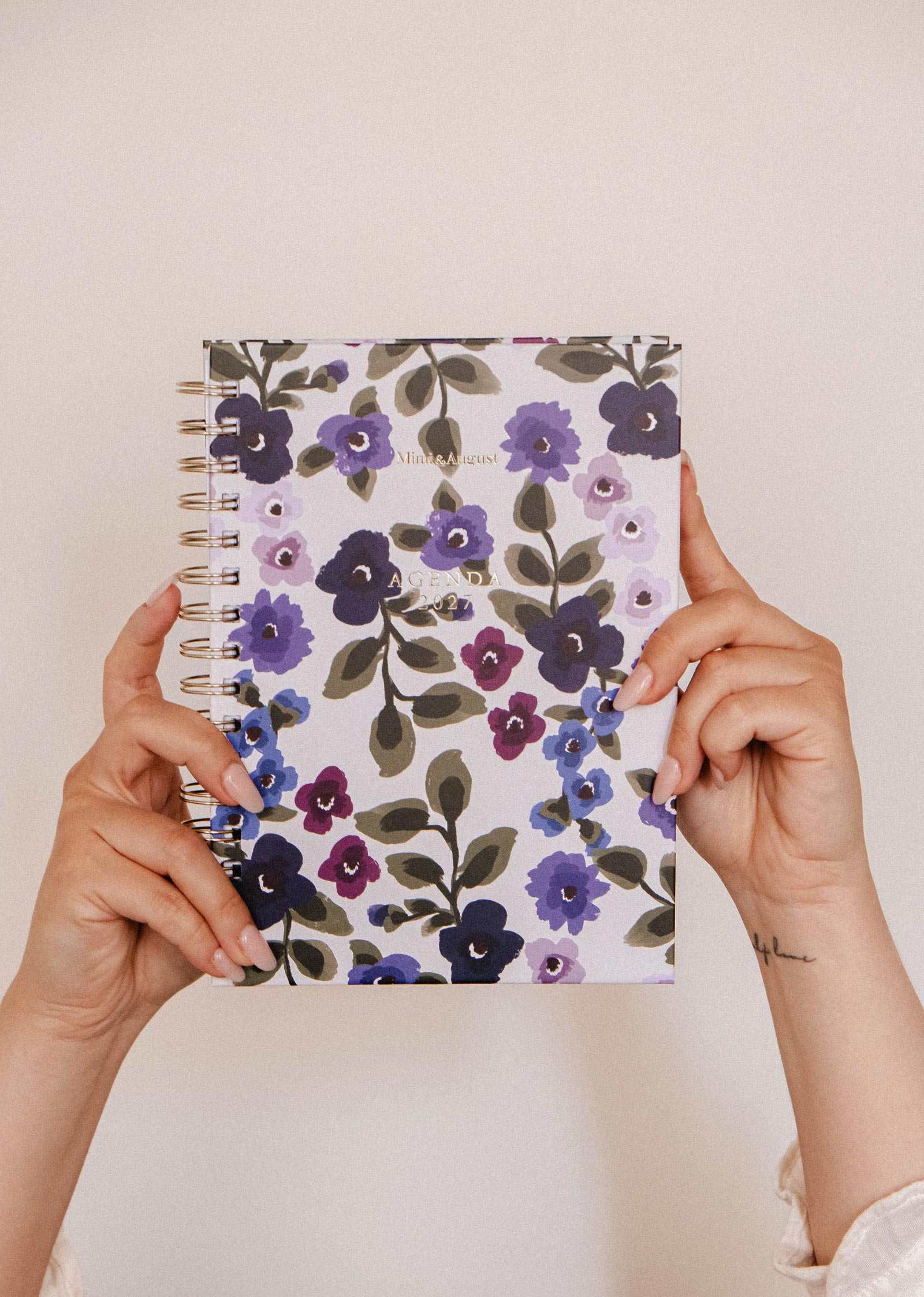 Two hands hold up the Mimi & August Wildflowers – 2027 Planner, featuring a spiral-bound notebook with a purple, lavender, and green floral cover against a plain beige background—perfect for monthly and weekly planning.