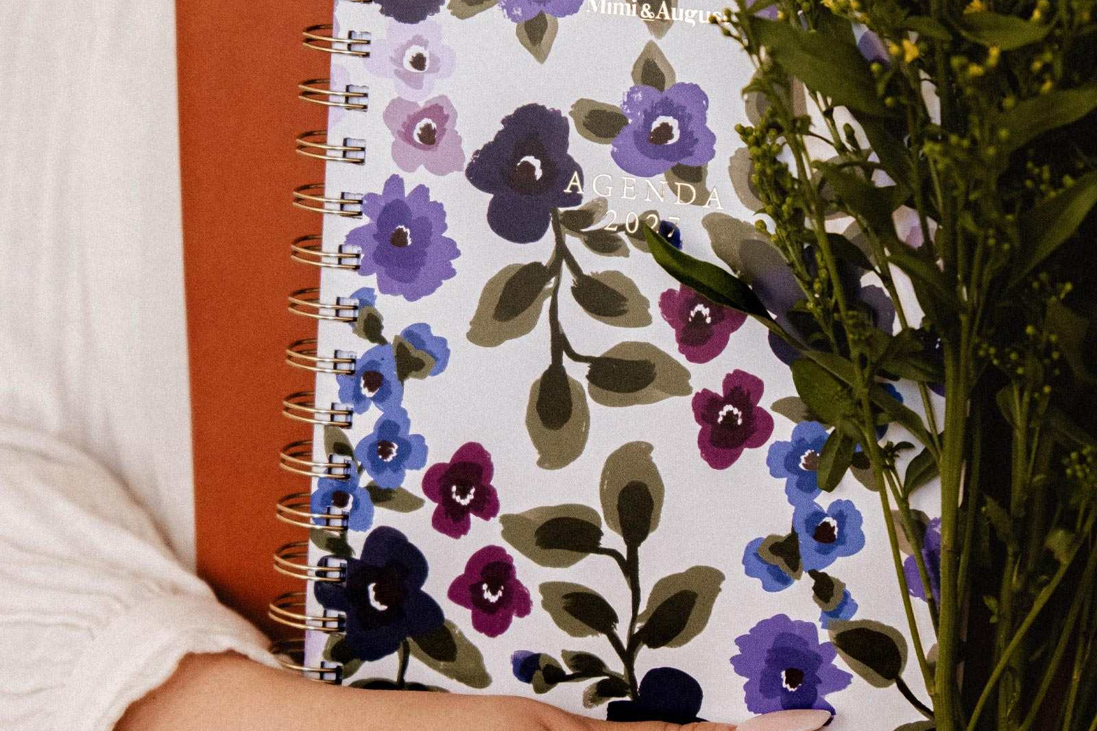 Une personne tenant un porte-documents brun, l'agenda Wildflowers 2027 de Mimi & August, un carnet spirale floral et un bouquet de fleurs jaunes, vêtue d'une chemise claire à manches longues