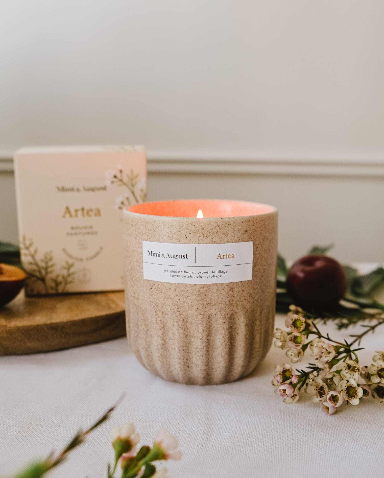 A beige floral Mimi & August Artea – Reusable Soy Candle is lit on a table with small flowers, greenery, and its box in the background, adding charm and sustainability to any space.