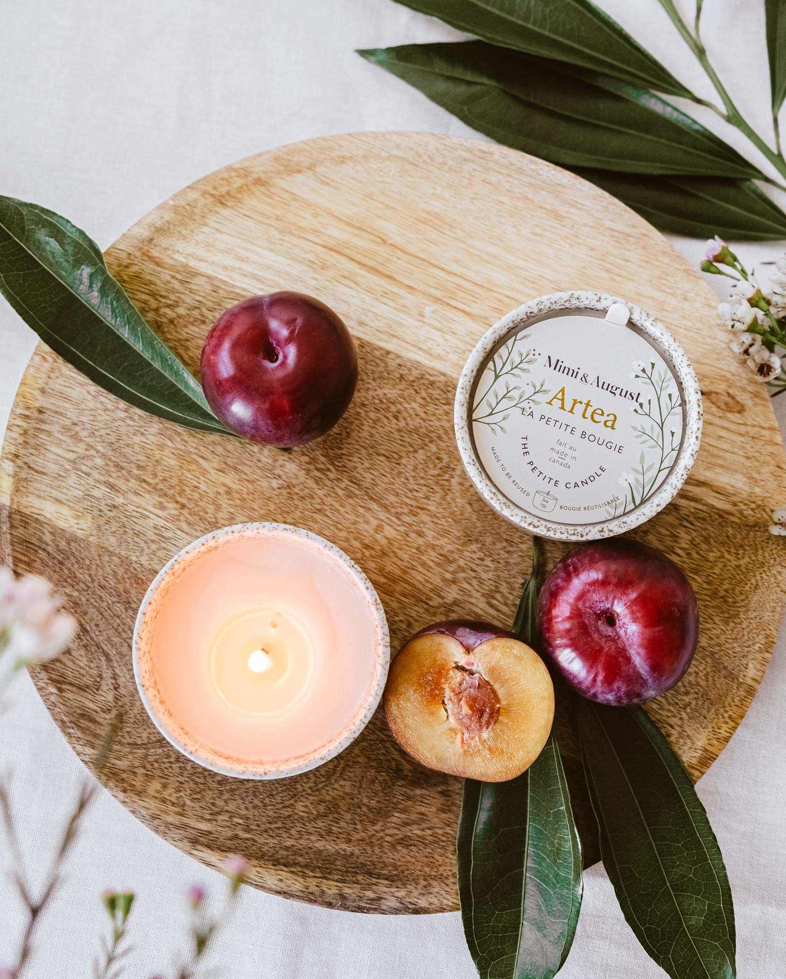 A lit Mimi & August Artea – Reusable Soy Candle in a ceramic container, plums, a halved plum, and the candle’s tin are arranged on a round wooden board with green leaves.