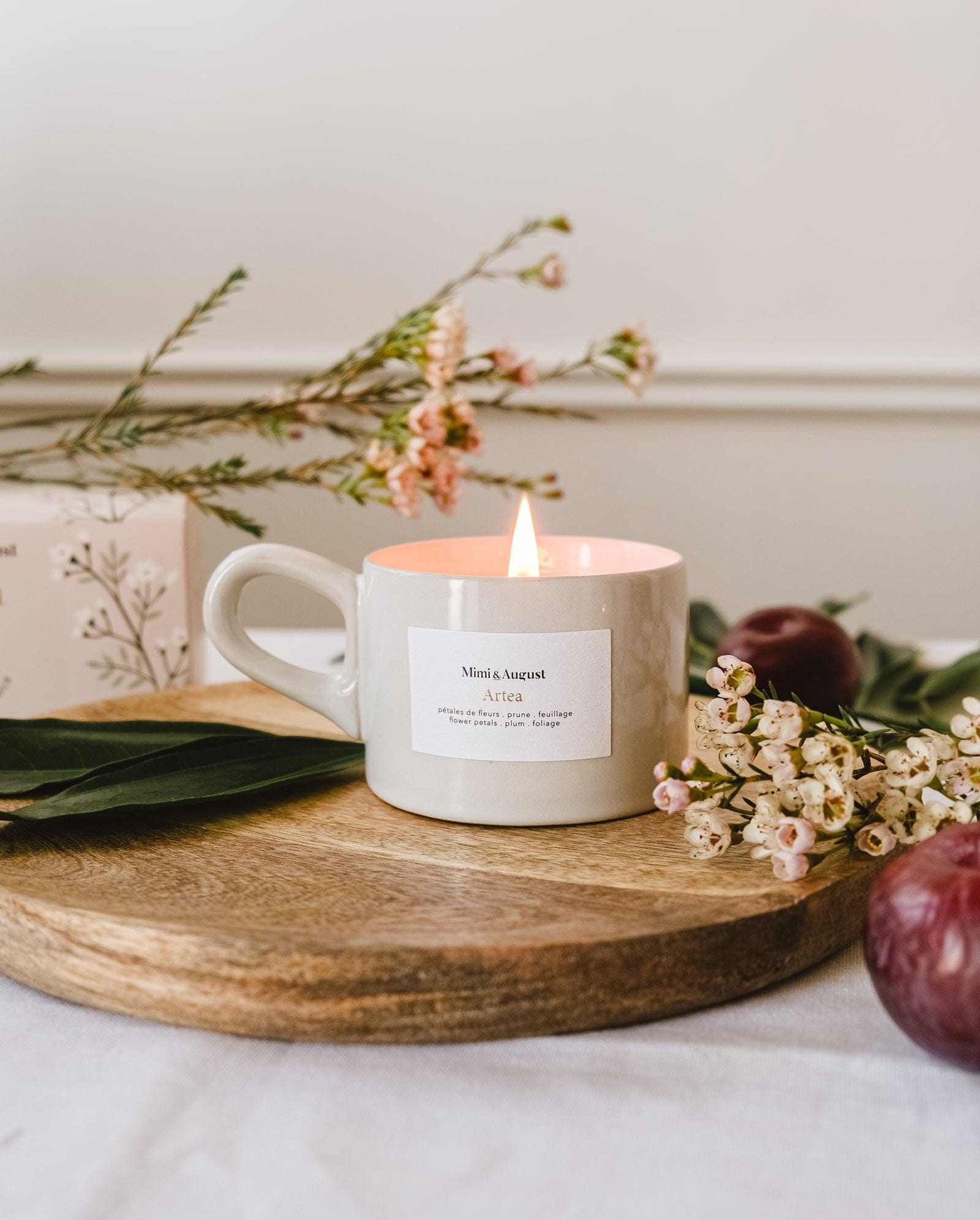 The Artea – Reusable Soy Candle by Mimi & August glows in a ceramic holder atop a wooden tray, surrounded by delicate flowers and leaves, with a plum and a floral box in the background.