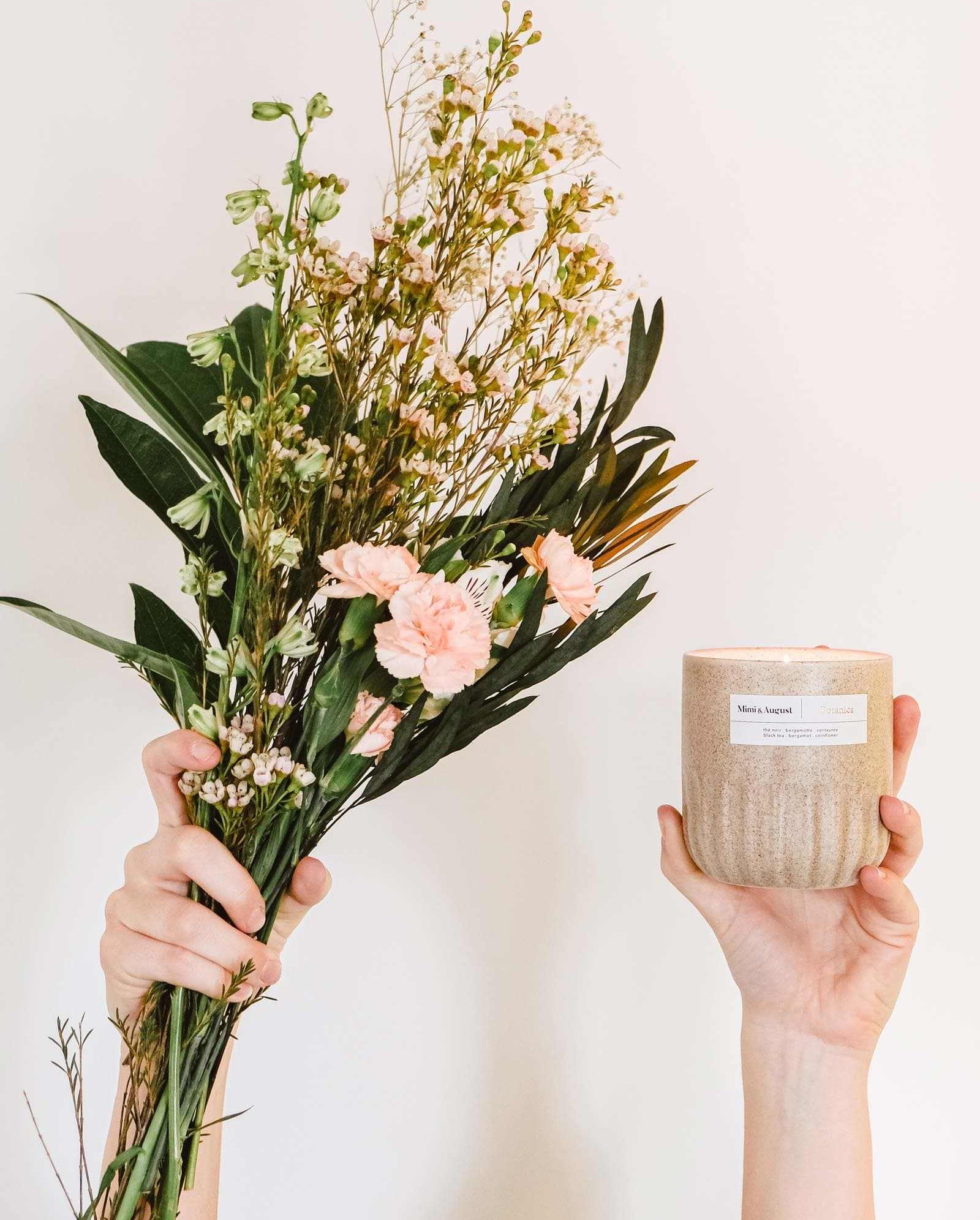 Hands holding a bouquet of pink flowers and a Mimi & August Botanica - Reusable Candle against a plain background.