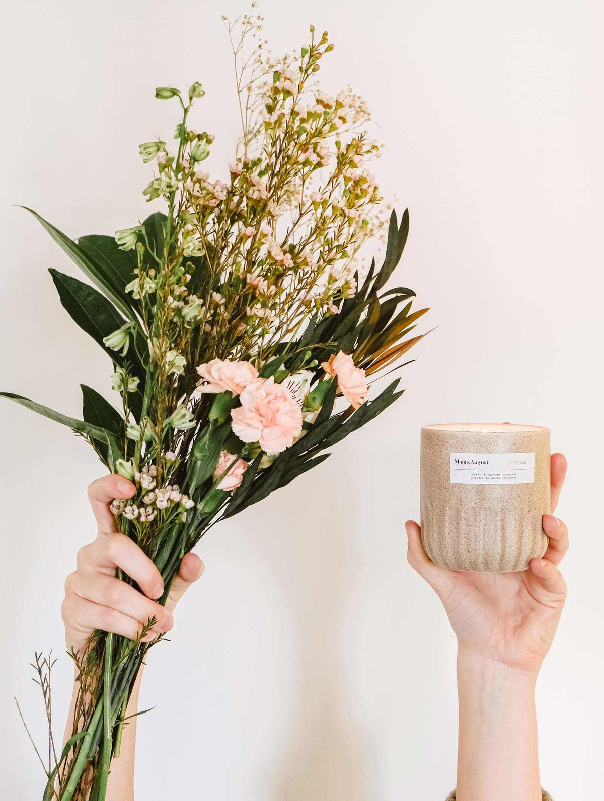 Hands holding a bouquet of pink flowers and a Mimi & August Botanica - Reusable Candle against a plain background.