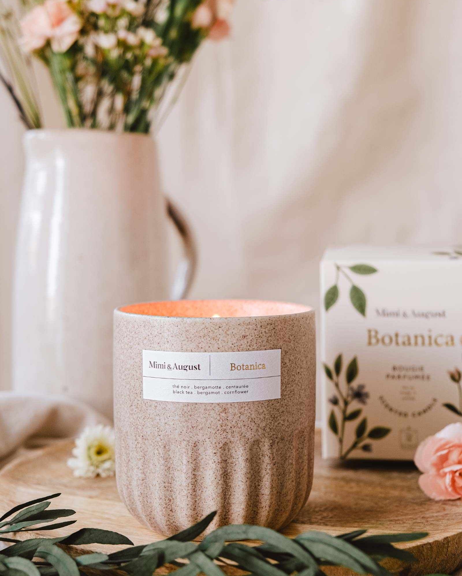 An aromatherapy Mimi & August Botanica - Reusable Candle on a wooden board with a floral arrangement and package in a cozy, homey setting.