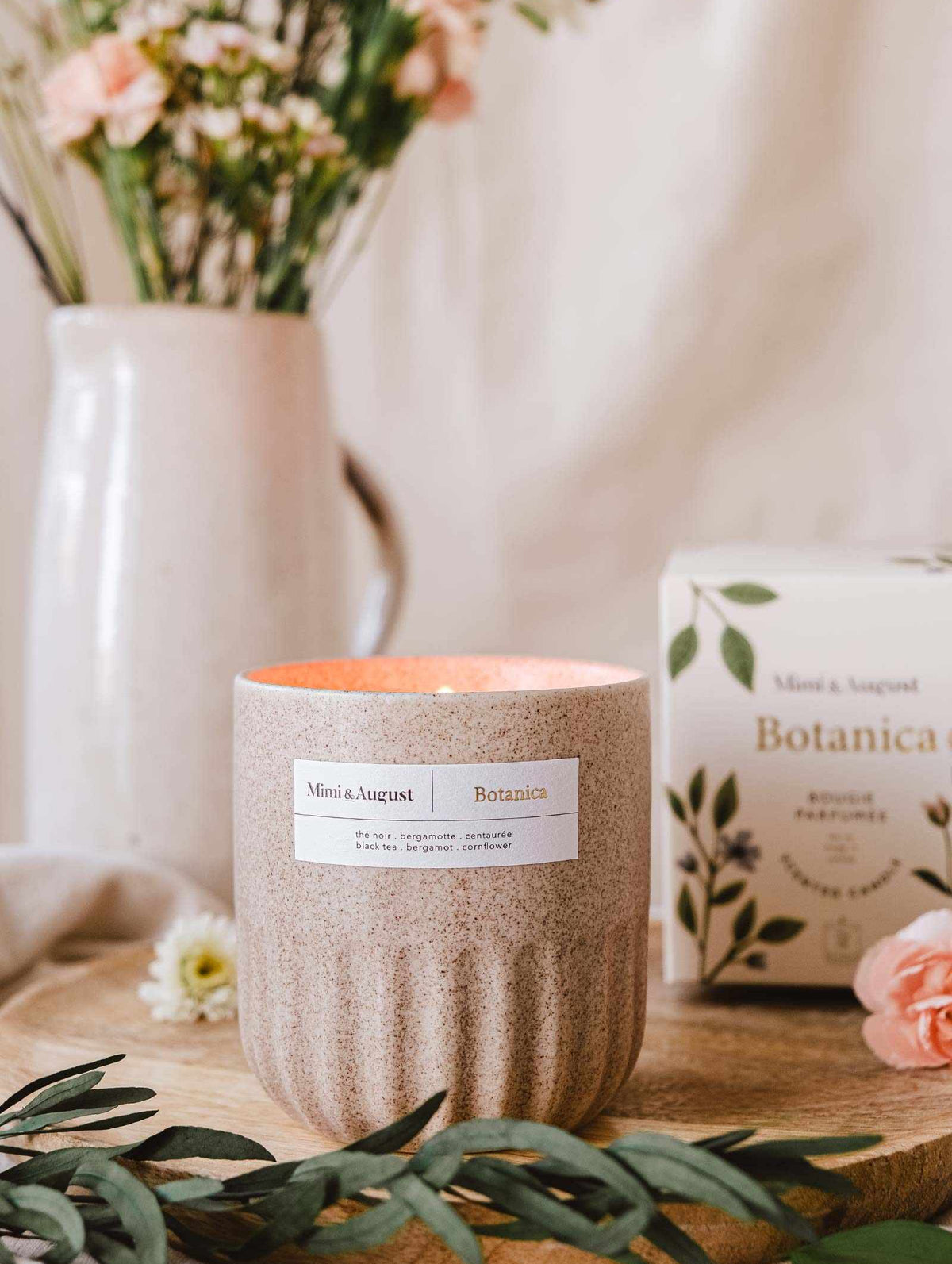 An aromatherapy Mimi & August Botanica - Reusable Candle on a wooden board with a floral arrangement and package in a cozy, homey setting.