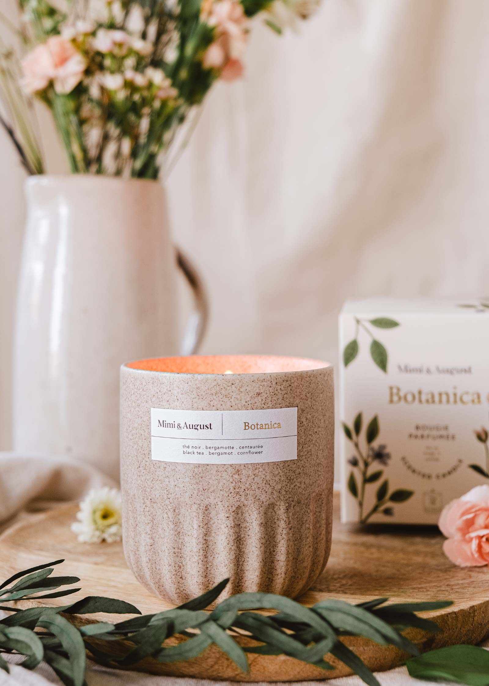 An aromatherapy Mimi & August Botanica - Reusable Candle on a wooden board with a floral arrangement and package in a cozy, homey setting.
