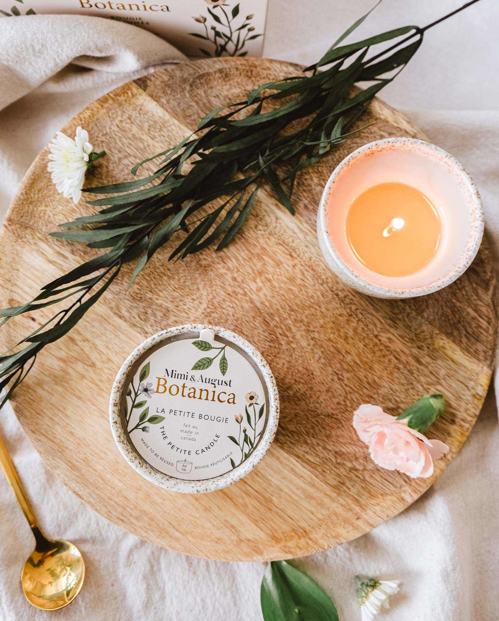 Flat lay of a wooden tray with a lit Mimi & August Botanica - Reusable Candle, beauty products, and scattered flowers, evoking a peaceful, spa-like atmosphere.