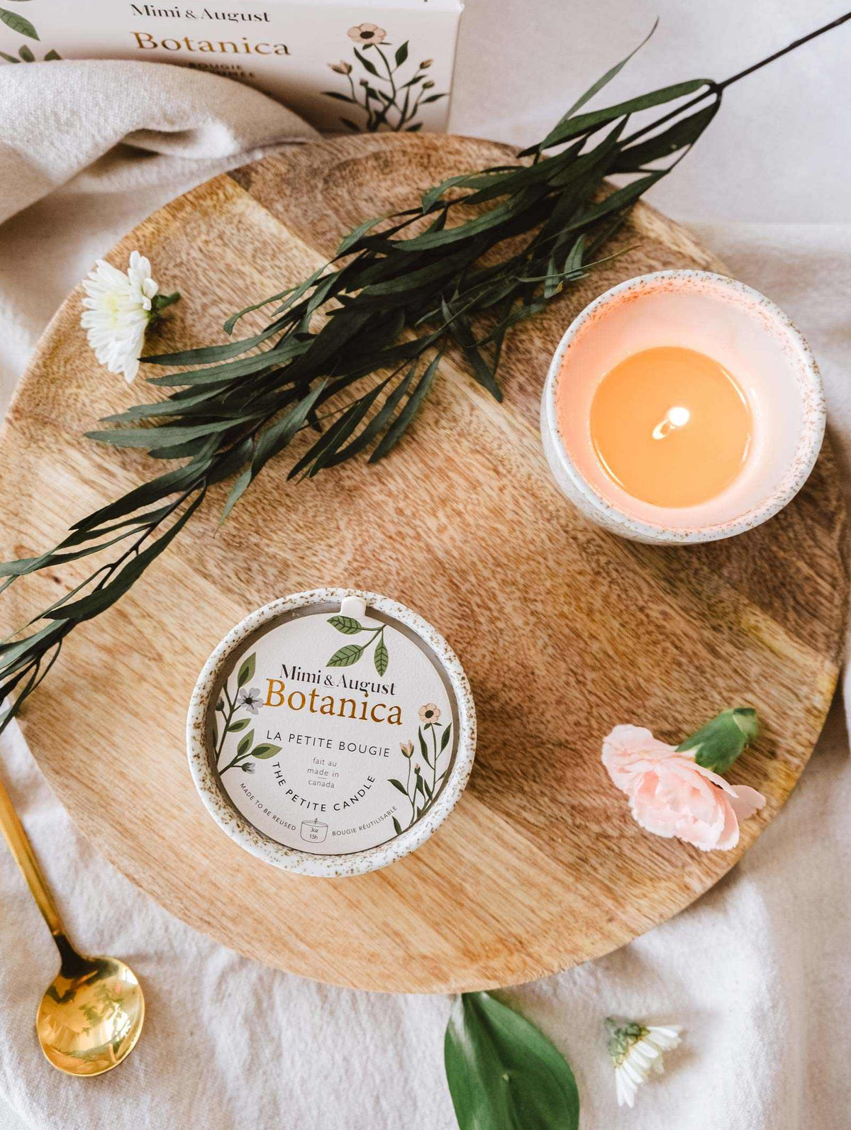 Flat lay of a wooden tray with a lit Mimi & August Botanica - Reusable Candle, beauty products, and scattered flowers, evoking a peaceful, spa-like atmosphere.