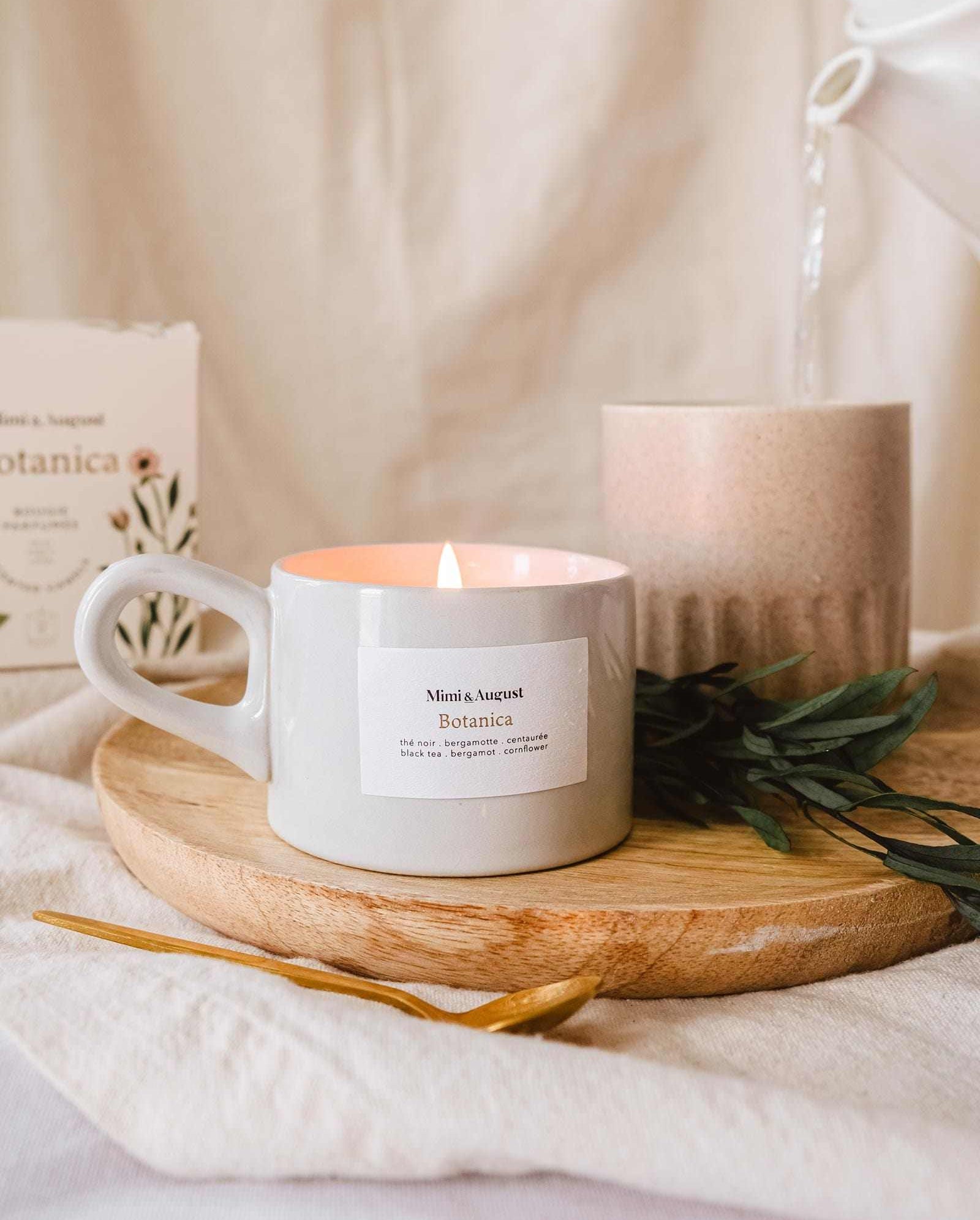 A lit Mimi & August Botanica - Reusable Candle on a wooden tray with a sprig of greenery and a decorative item in a cozy, softly lit setting.