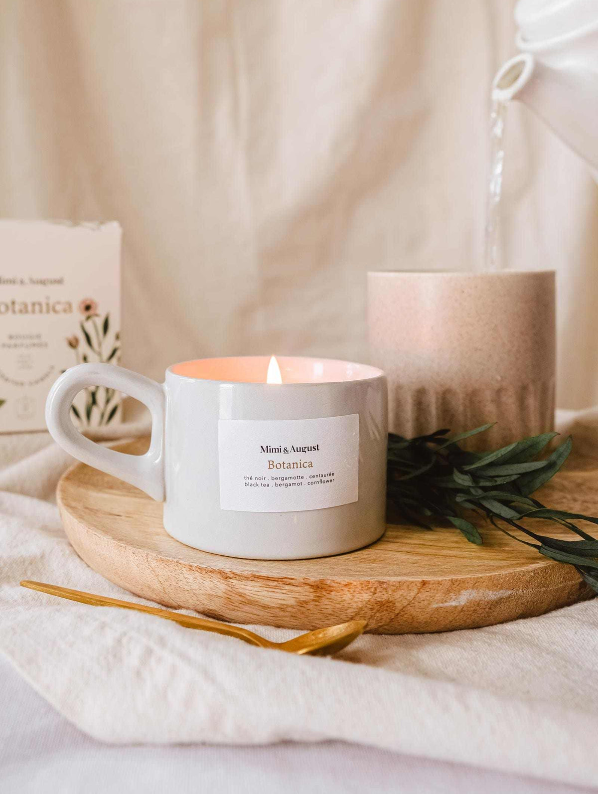 A lit Mimi & August Botanica - Reusable Candle on a wooden tray with a sprig of greenery and a decorative item in a cozy, softly lit setting.