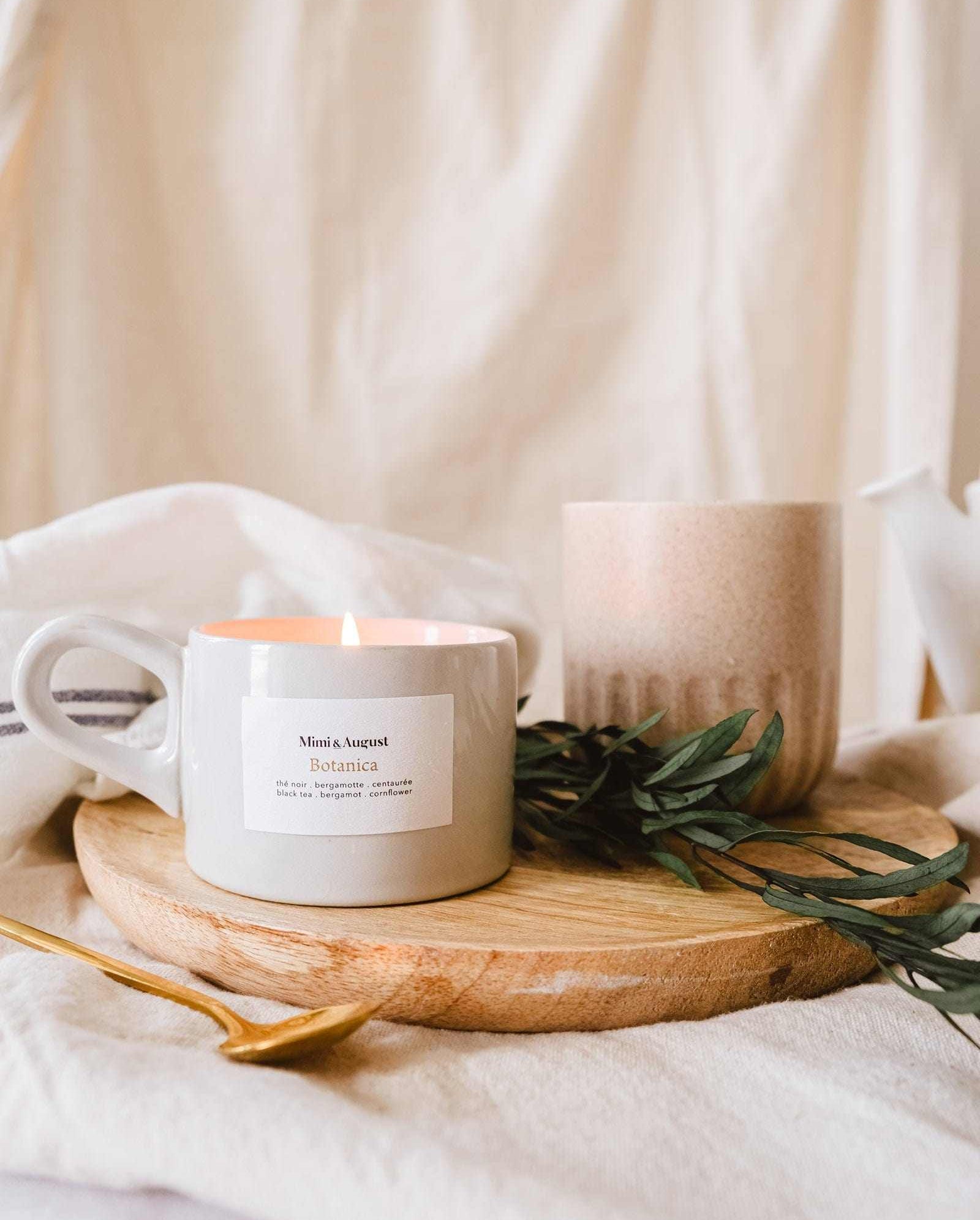 A lit Mimi & August Botanica - Reusable Candle on a wooden tray with green leaves, surrounded by a ceramic mug and soft beige textiles.