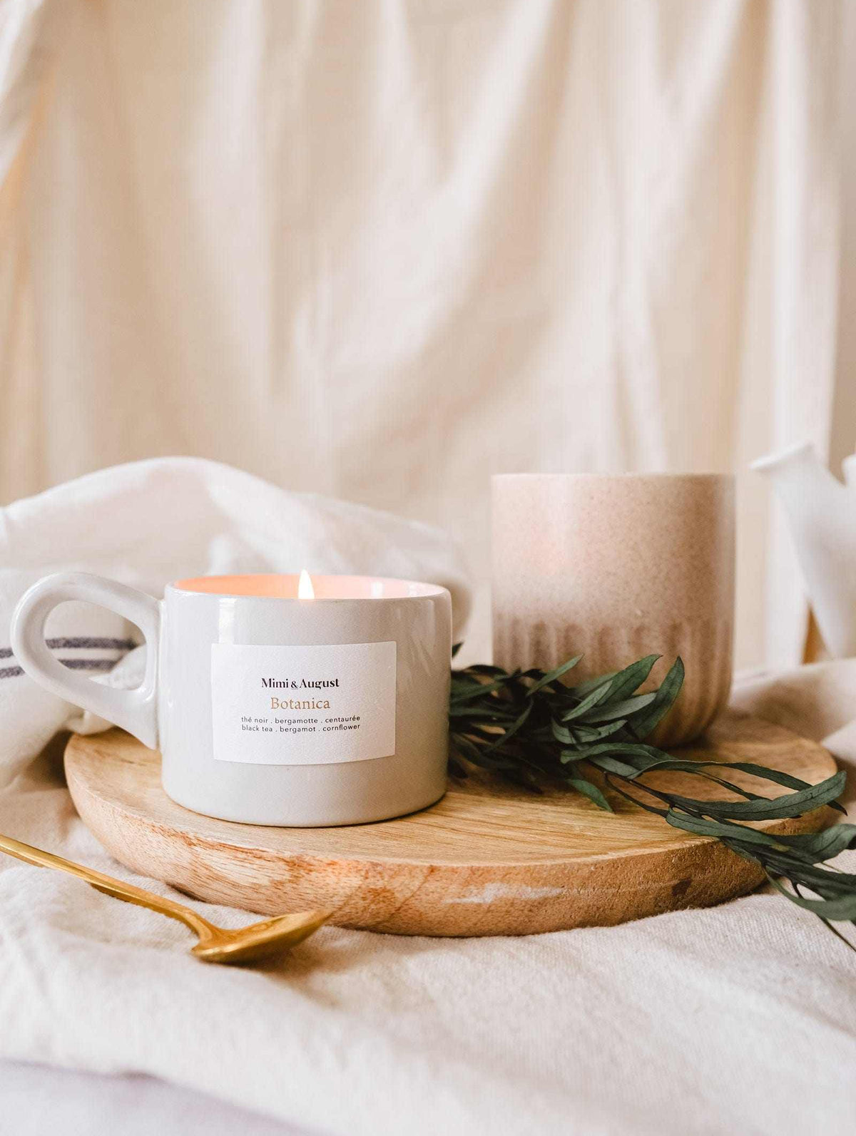 A lit Mimi & August Botanica - Reusable Candle on a wooden tray with green leaves, surrounded by a ceramic mug and soft beige textiles.