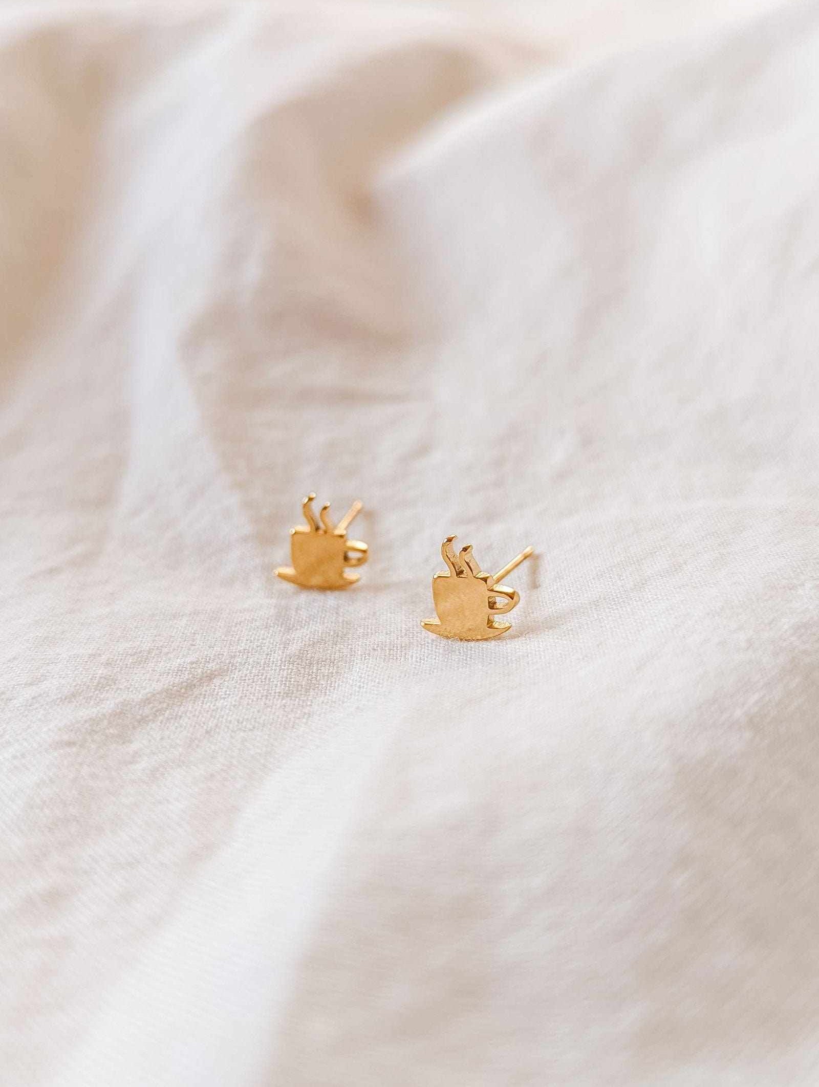 Cafecito gold plated earrings shaped like coffee cups on beige fabric background