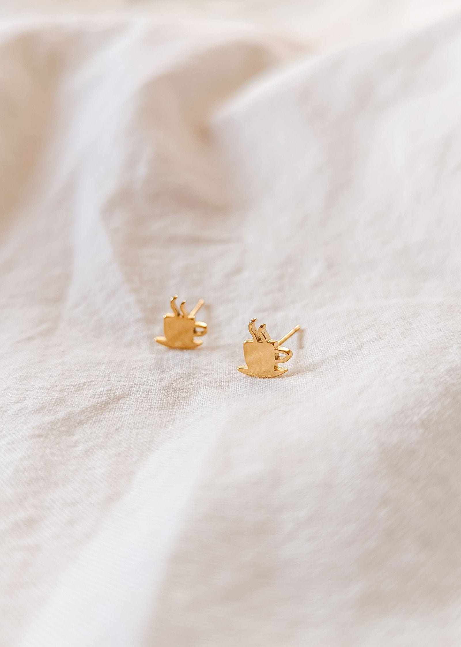 Cafecito gold plated earrings shaped like coffee cups on beige fabric background