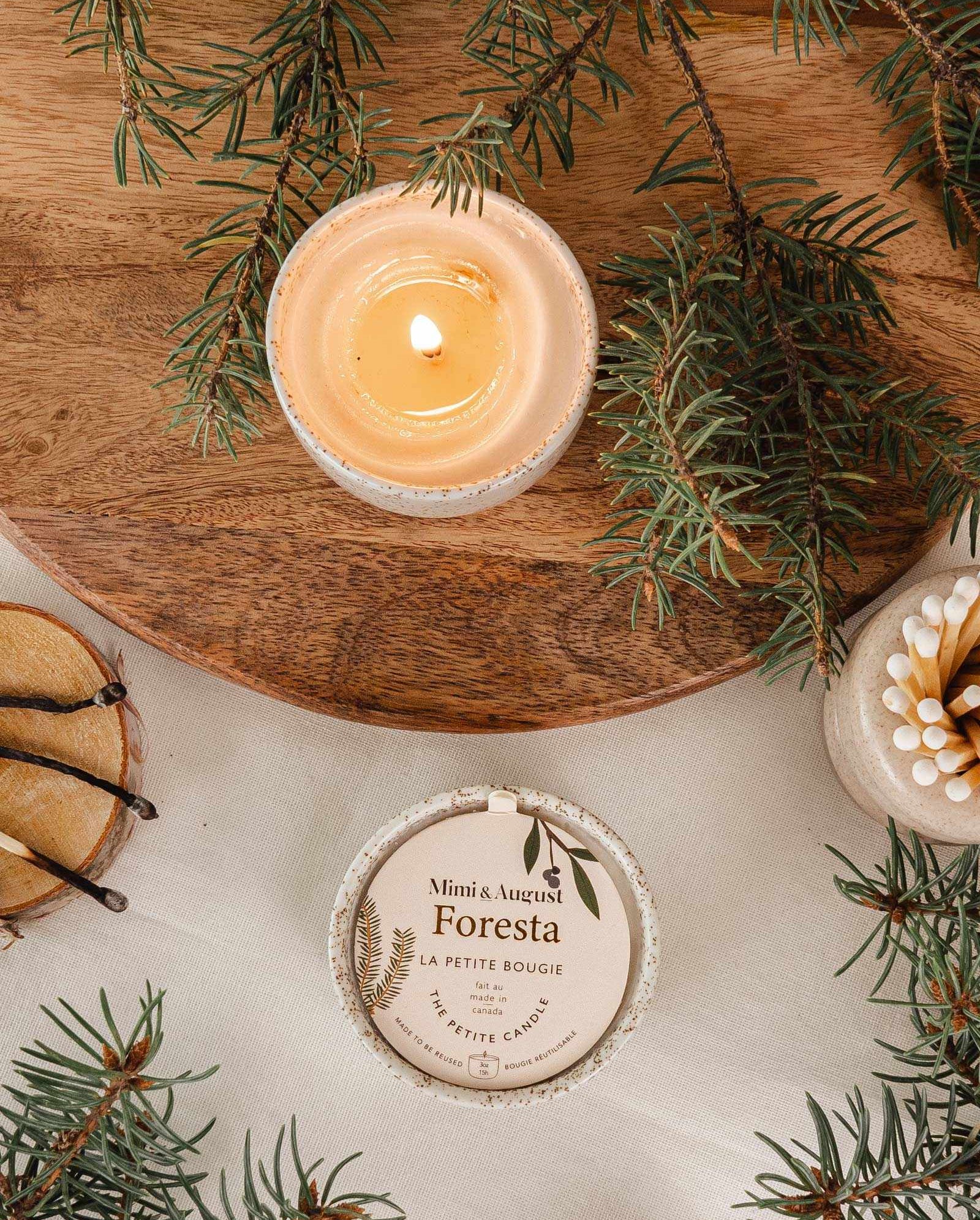 Lit fir tree candle in reusable ceramic cup surrounded by fir branches and candle lid on wooden surface