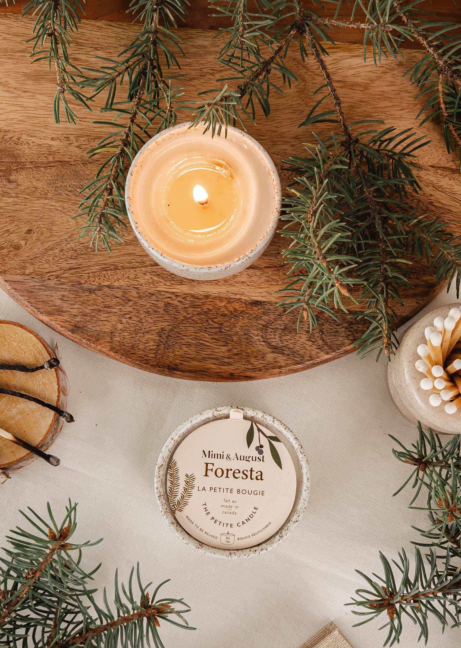 Lit fir tree candle in reusable ceramic cup surrounded by fir branches and candle lid on wooden surface