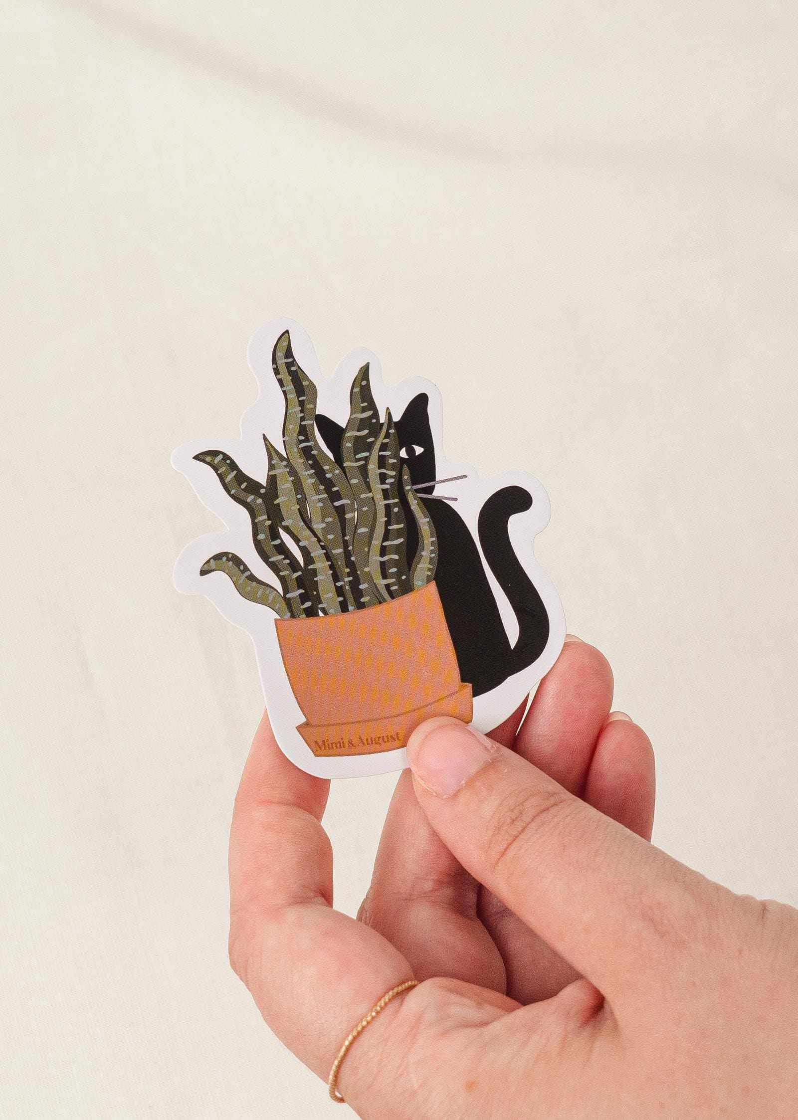 Hand holding durable vinyl sticker of black cat peeking from behind a potted plant with white border