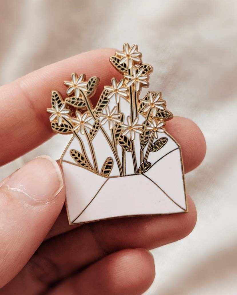 Hand holding a 38mm gold tone brass enamel pin shaped like an envelope filled with flowers, titled Letter full of flowers.