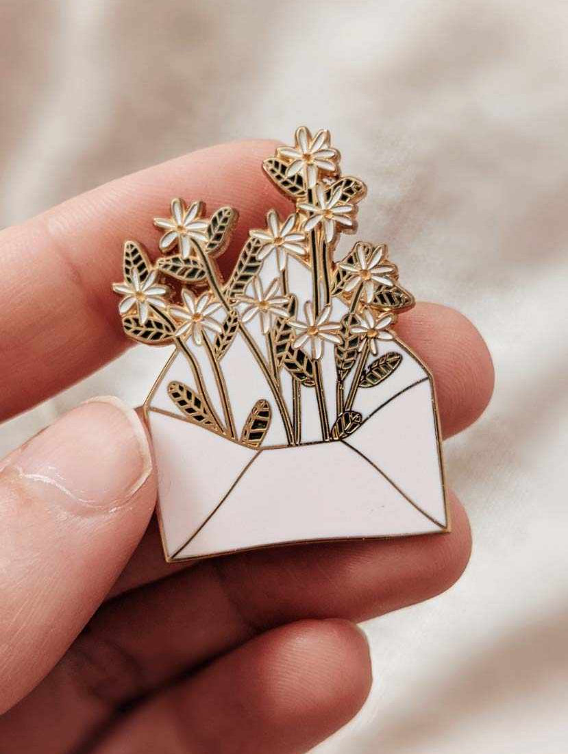 Hand holding a 38mm gold tone brass enamel pin shaped like an envelope filled with flowers, titled Letter full of flowers.