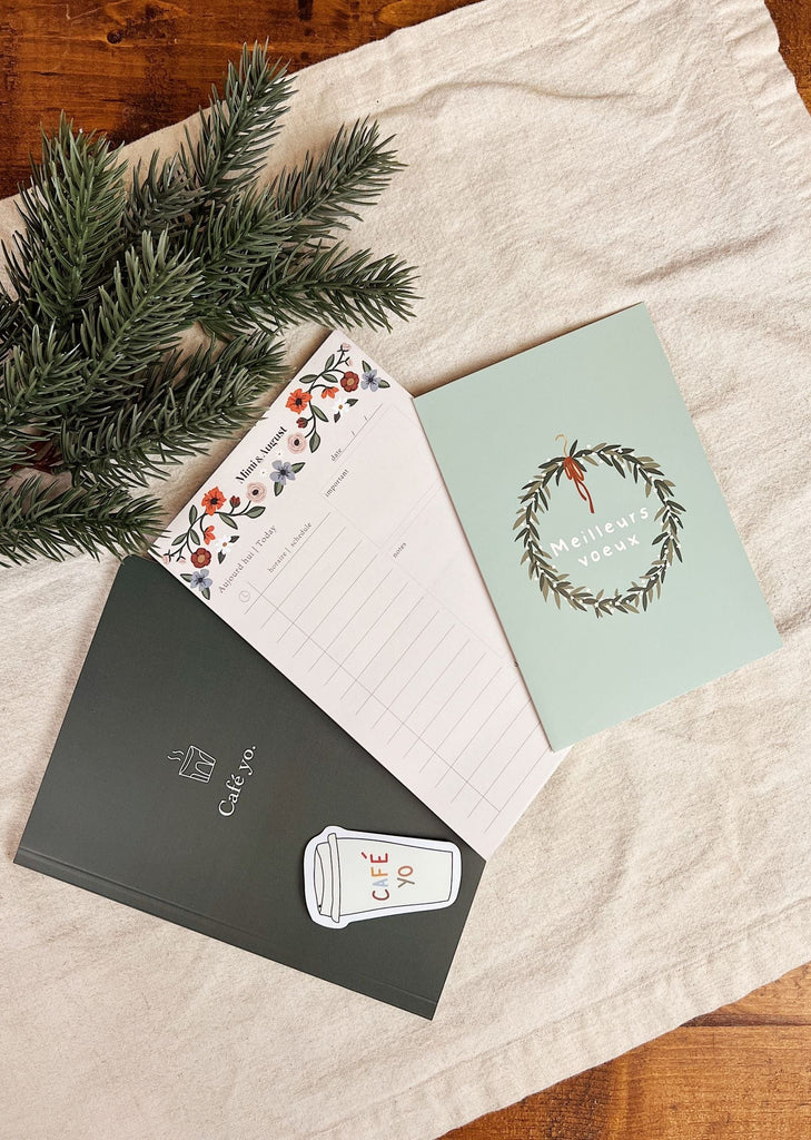 A The little elf planner bundle from Mimi & August, two festive Meilleurs voeux greeting cards, a pen, and a branch of greenery are arranged on a cloth-covered surface.