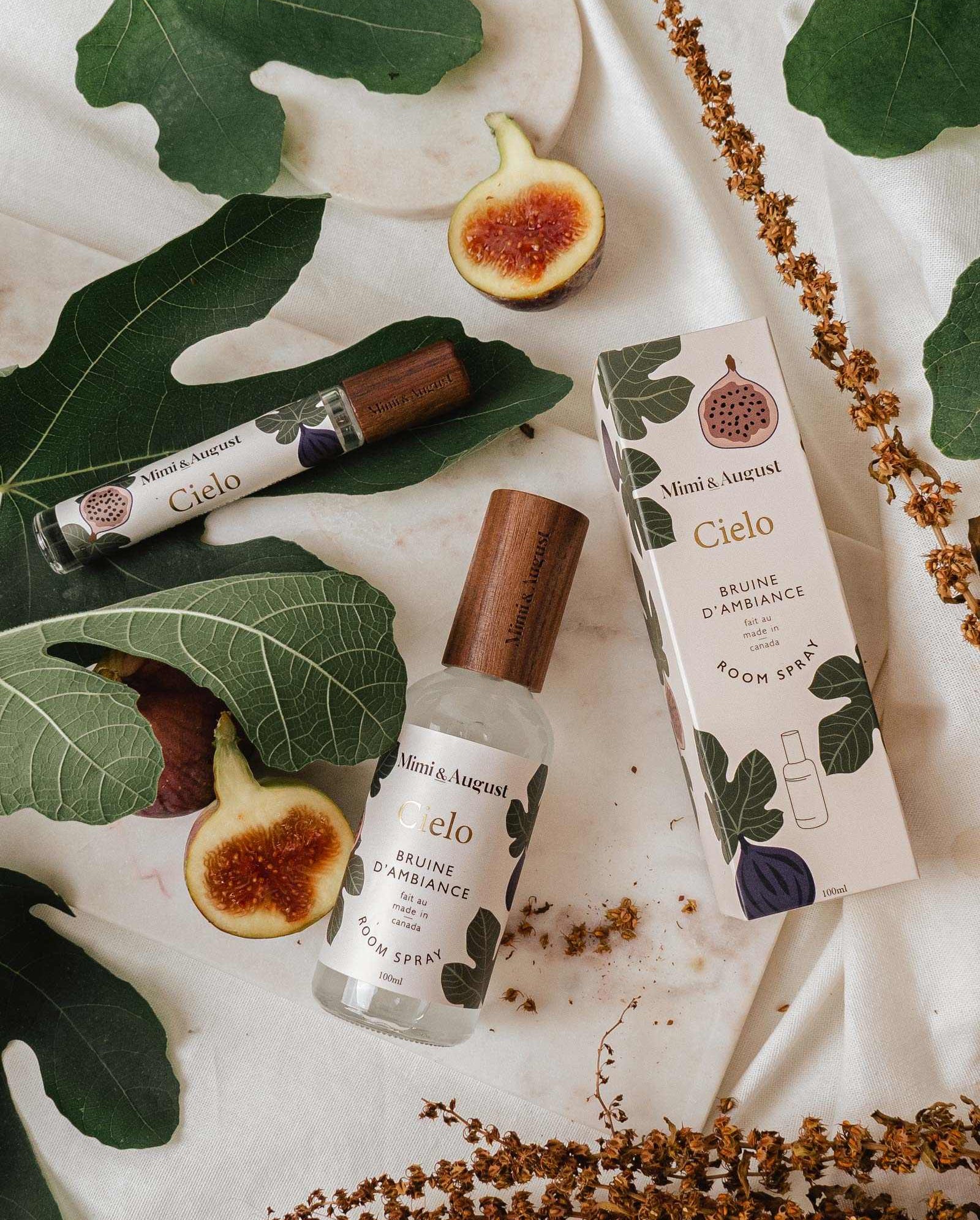 A bottle and box of Mimi & August Cielo Room Spray, made with premium fragrance oil, are displayed on a white cloth decorated with fig leaves, dried flowers, and fresh figs.