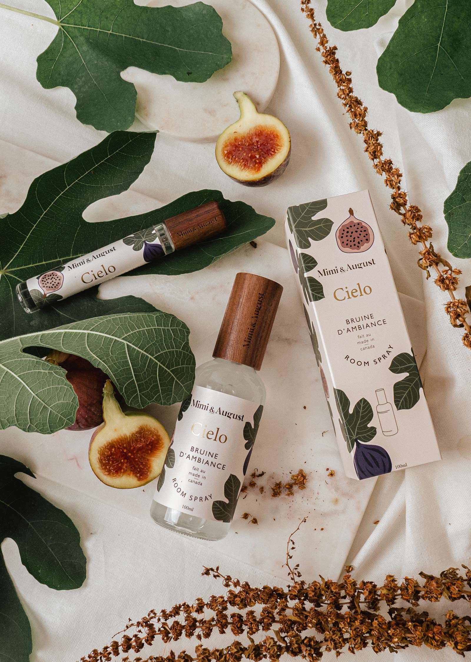 A bottle and box of Mimi & August Cielo fig room spray, made with premium fragrance oil, are displayed on a white cloth decorated with fig leaves, dried flowers, and fresh figs.