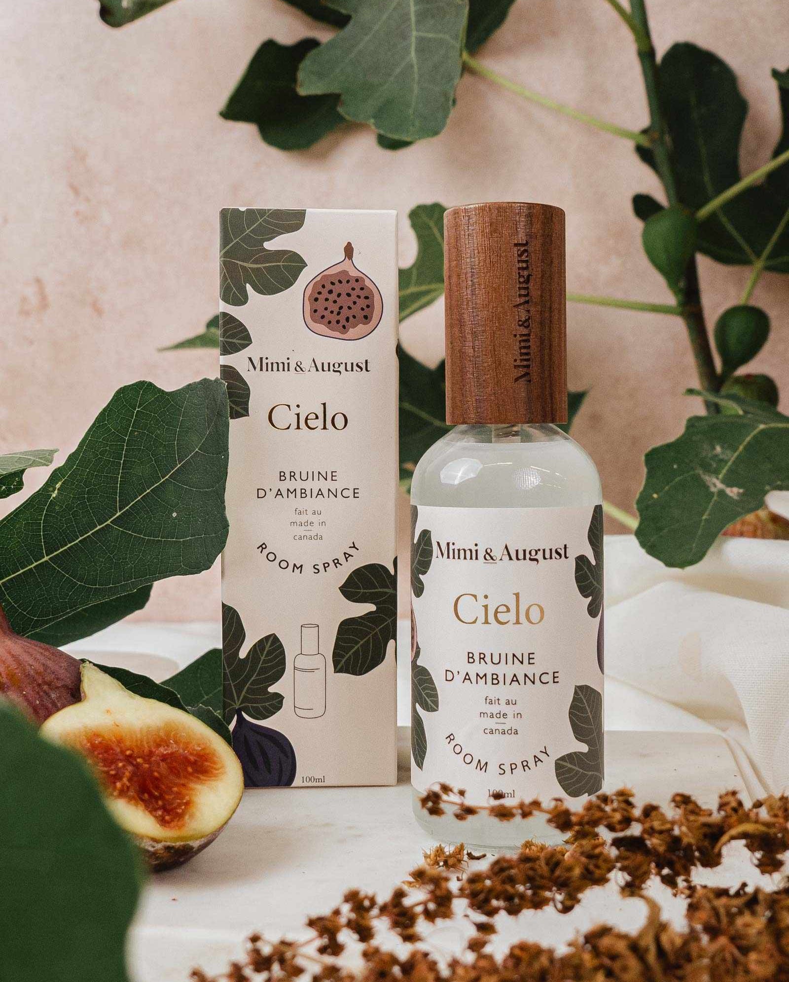 A Mimi & August Cielo Room Spray bottle and box sit on a surface with fig leaves, a halved fig, and dried flowers in the background. Crafted with premium fragrance oil for an elevated scent experience.