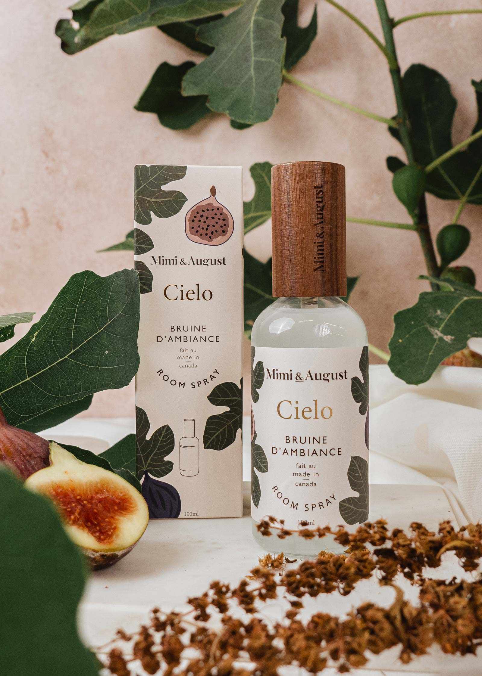 A Mimi & August Cielo fig room spray bottle and box sit on a surface with fig leaves, a halved fig, and dried flowers in the background. Crafted with premium fragrance oil for an elevated scent experience.